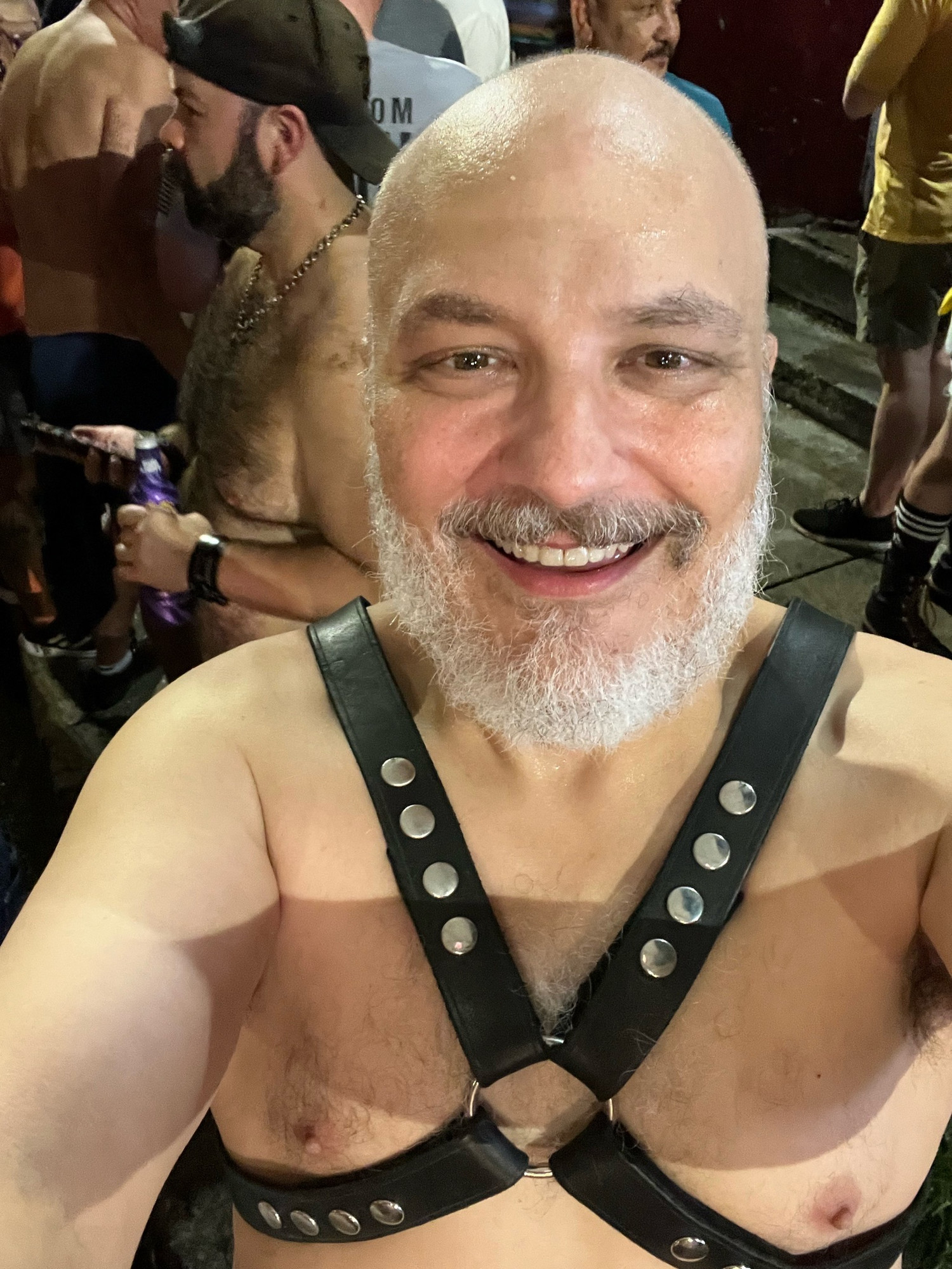 Bald man in leather harness smiles at camera while standing on street outside gay bar.