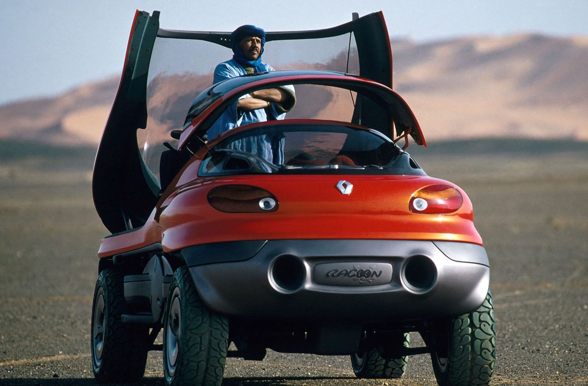 Photo of the Raccoon in a desert, shot from behind. A man is standing up in the front seat with the hatch open, spaceship-style, facing backward.