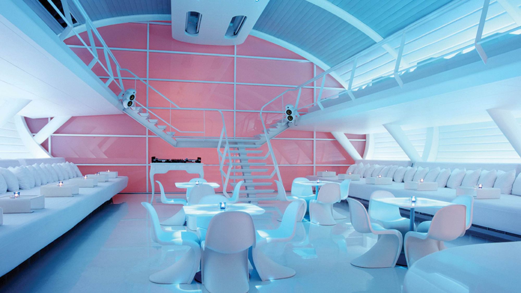 Interior of a futuristic restaurant shaped like a large oval tube. Spaceship-esque balconies and a floating staircase descend to a dining area edged by two extremely wide beds. In the center, swoopy molded chairs sit at sleek plastic tables. Everything is white and pale blue, except for the large frosted window comprising the far wall, which glows a warm pink.