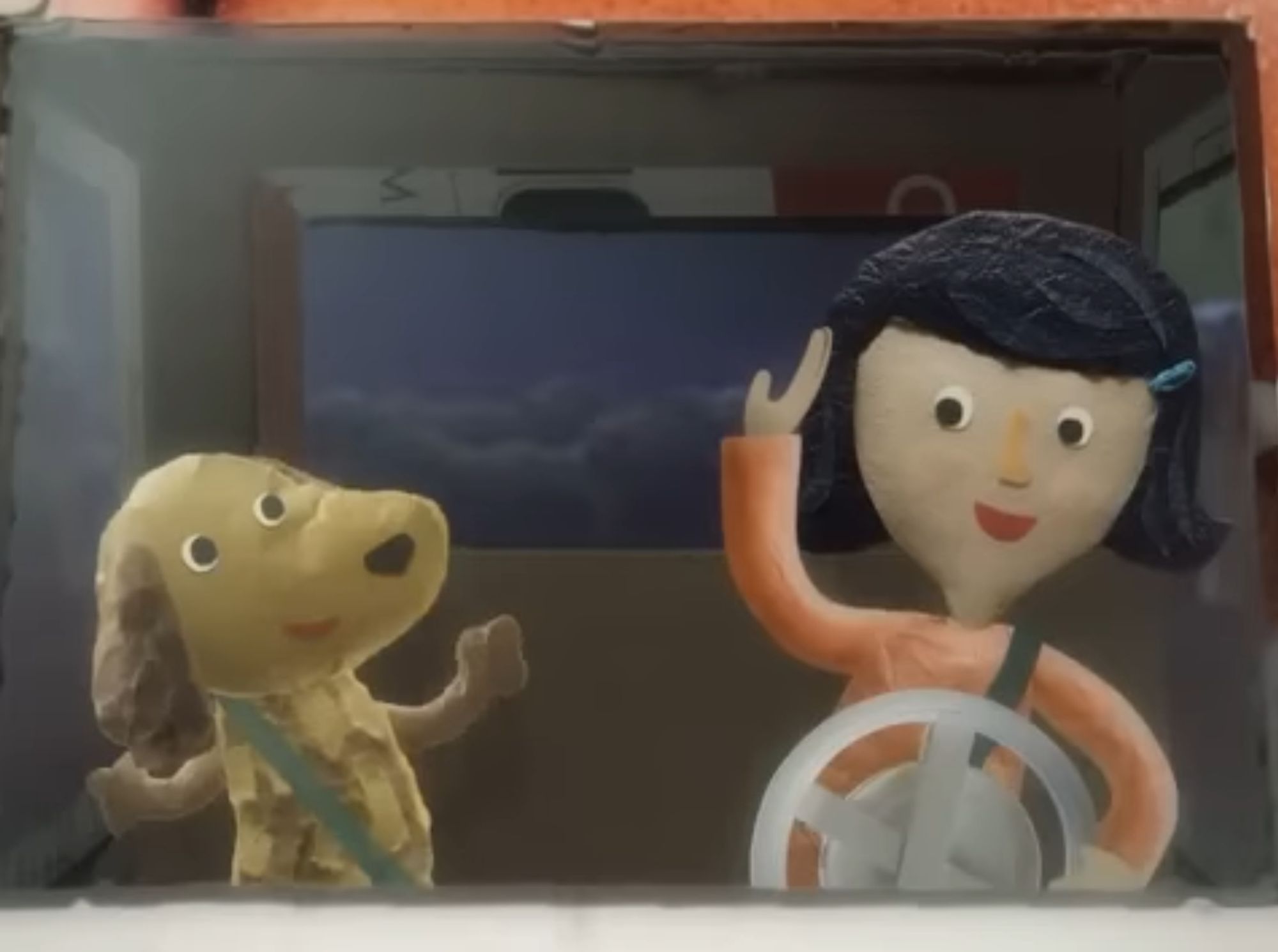 Still from the music video. A woman drives the titular electric car, which is made of cardboard and cut-up cereal boxes, and is just frames away from hi-5ing a smiling dog in the passenger seat.