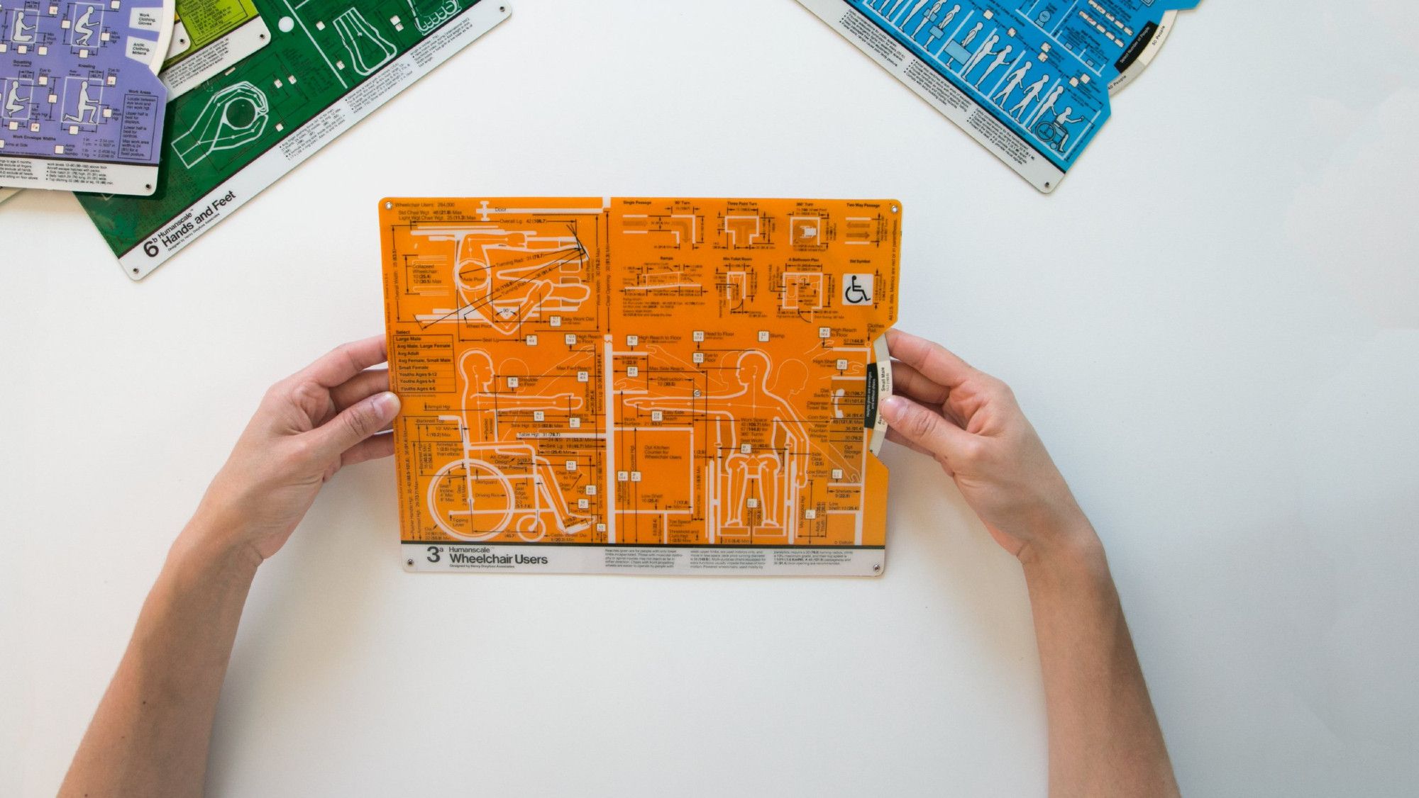 Hands holding a reference card from the guide, a diagram of a wheelchair user's proportions and measurements printed in black and white on a bright orange background at maximum density.