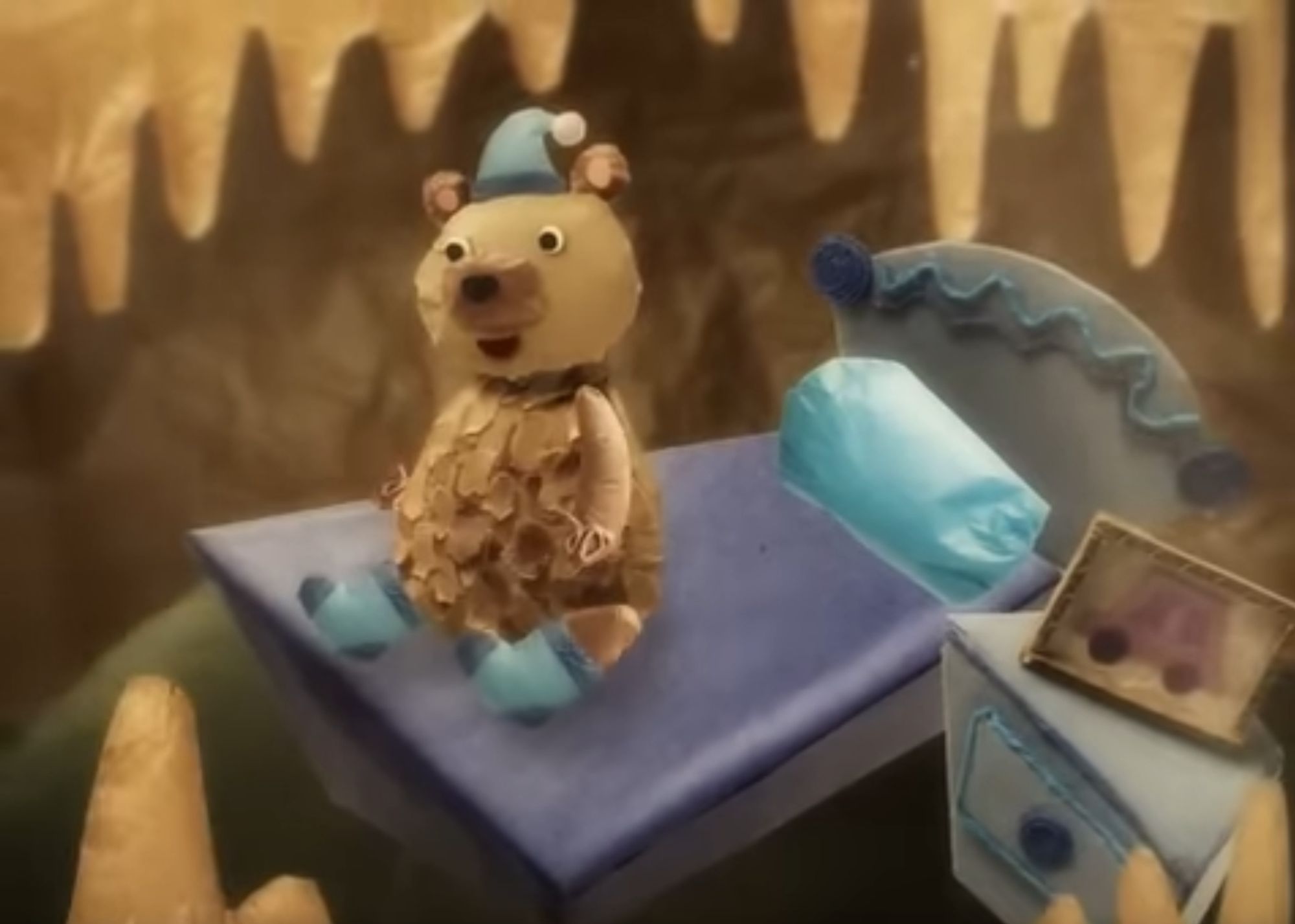 Still from the video of a smiling bear, made out of shredded brown paper, waking up in a purple bed in a paper-mache cave. On his nightstand is a framed picture of the electric car.