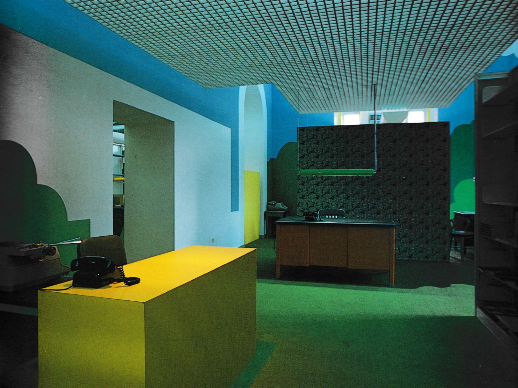 A wider shot of the same room. Playful colors and abstract graphics fill the space around two desks, one a standard oak-veneer affair and the other a geometric yellow rectangle devoid of features.