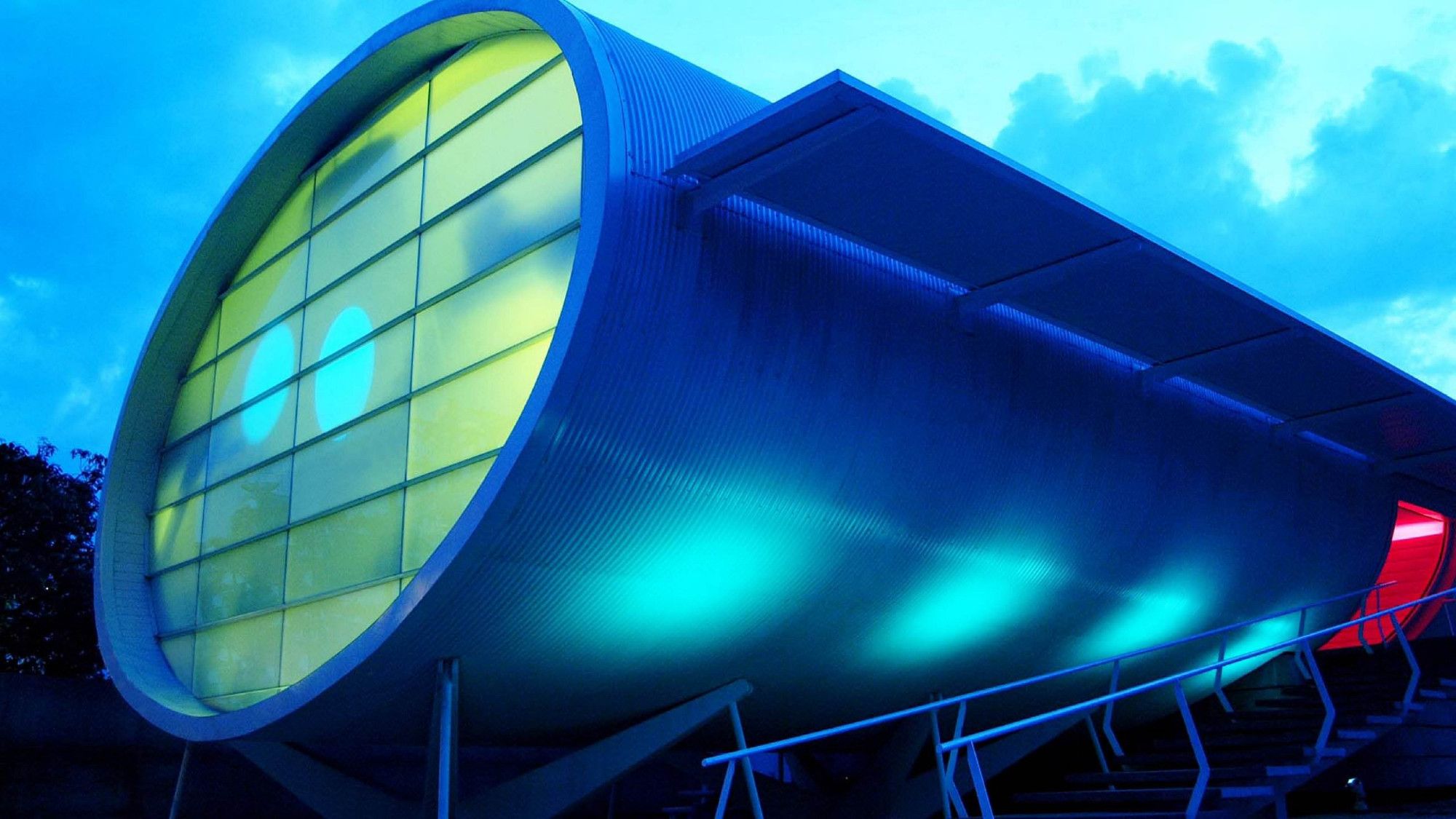 An exterior photo of the same building. It's a large corrugated metal cylinder resting on its side on an X-shaped support structure. A flat, modernist awning shades a long staircase which leads from the front of the tube--the side with the pink window--to a UFO-style convex doorway on the rear right side of the tube.