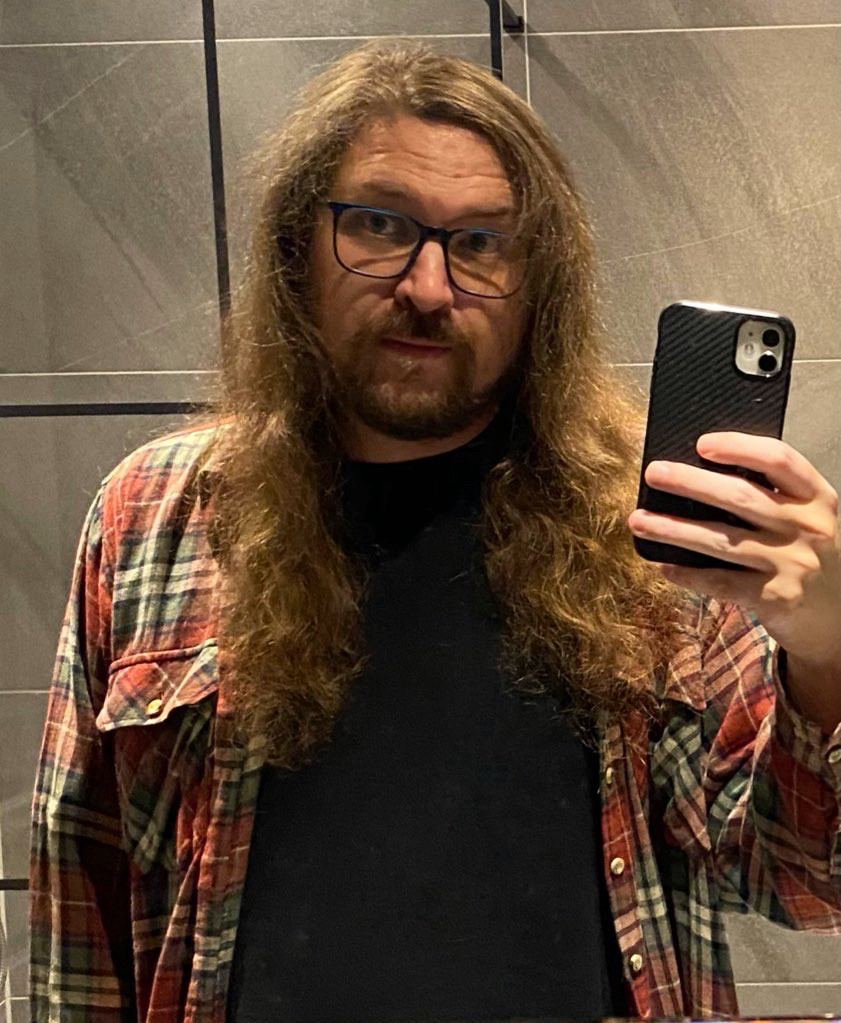 Caucasian male with light brown long hair and facial hair wearing glasses, a black T-shirt and plaid green and red shirt 