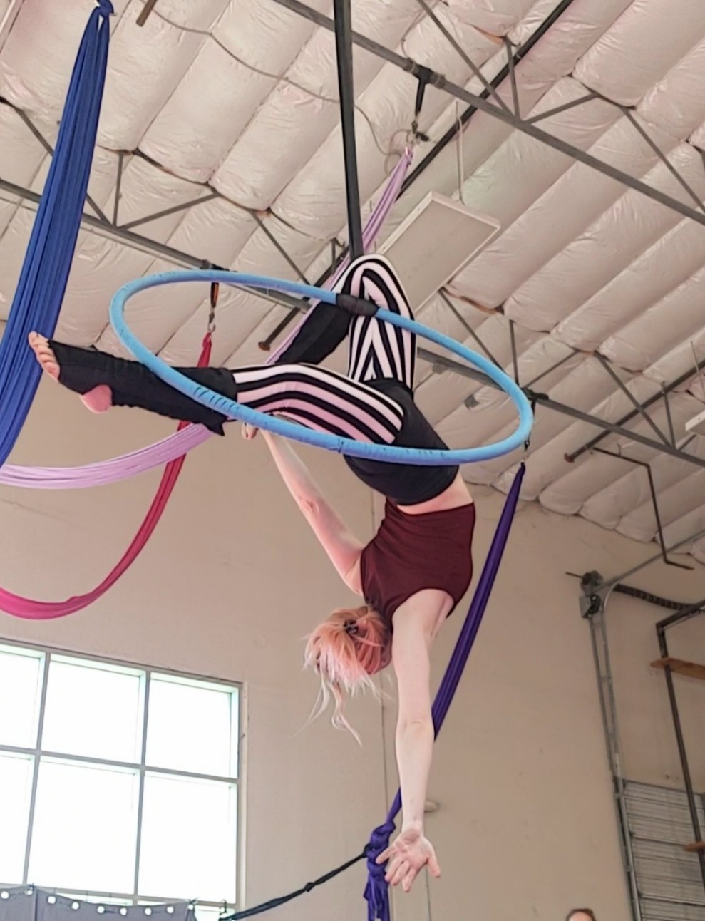 An aerialist hanging upside down, their right knee hookednon the spanset and the left leg pointed straight