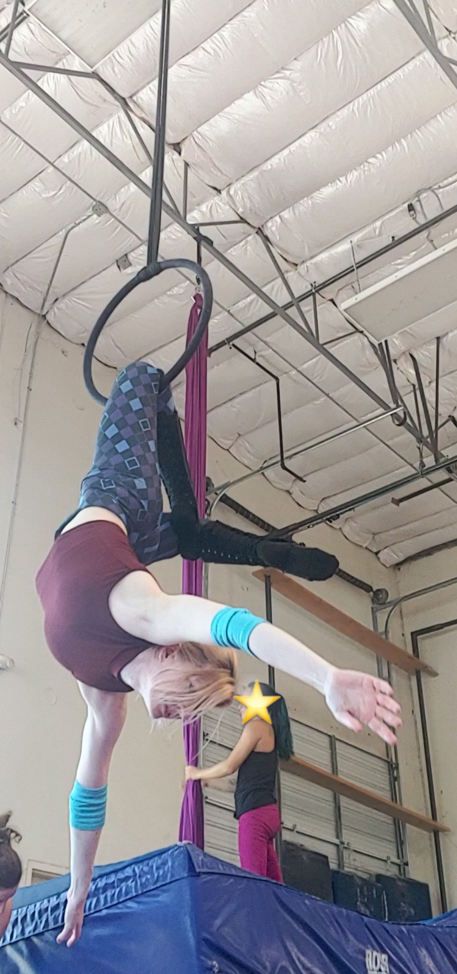 An aerialist hangs upside down from one knee on a mini-hoop. The foot of the hooked leg then rests behind the knee of the free leg. The arms hang free