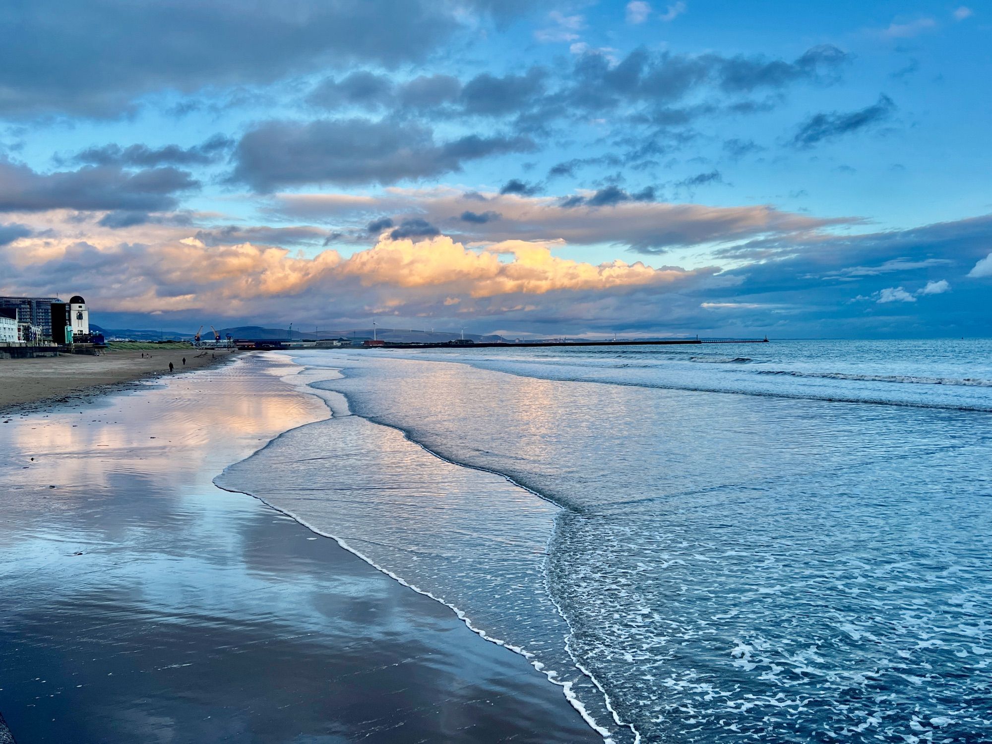 Early evening golden light on clouds in the eastern sky above Swansea Bay with a quiet tide sliding shallow over a reflective beach. All blues and greys and gold.