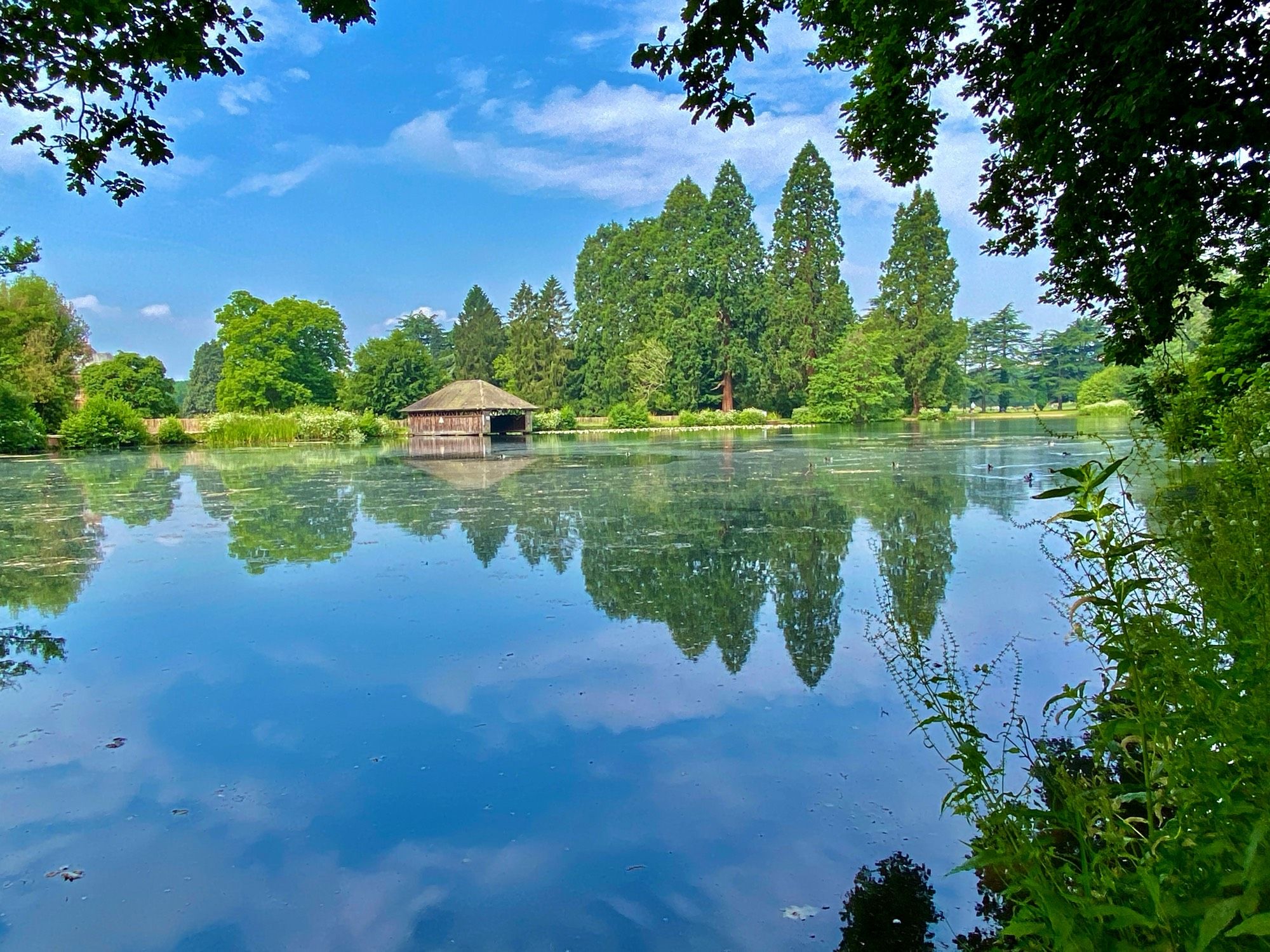 A view of the lake at Tredegar House, Newport, with a still calm lake reflecting the blue sky and trees and a wooden boat house across the water.