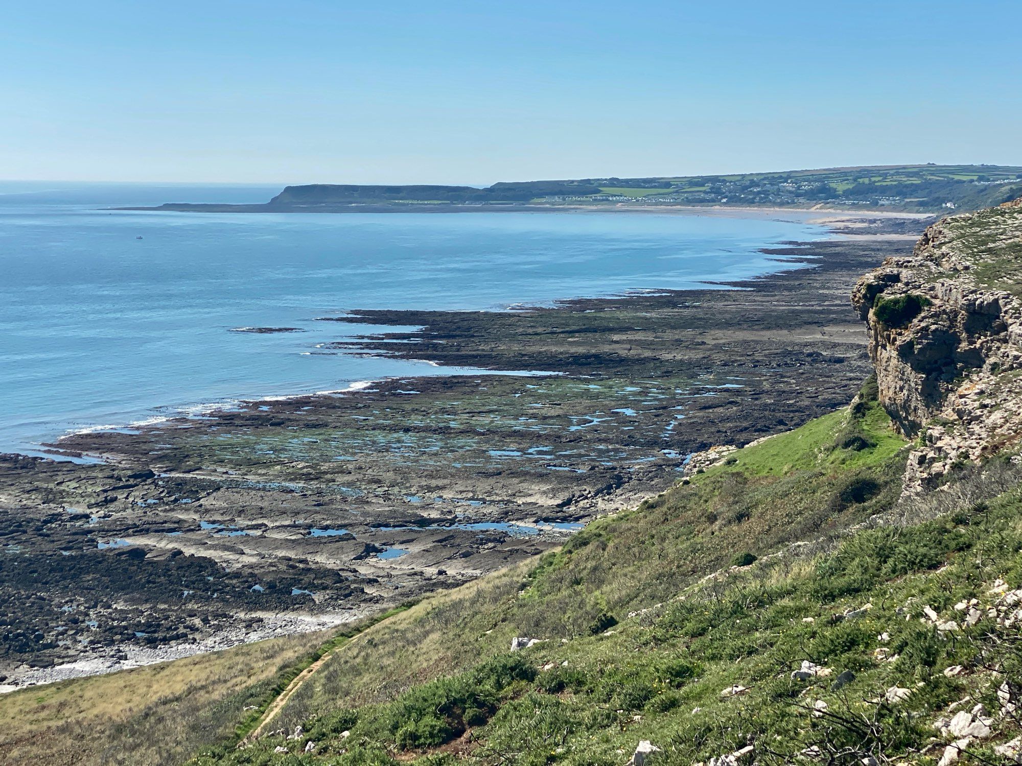 Low tide dark rocks stretching out with hundreds of rockpools reflecting the blue sky. Blue sea and sky with Oxwich Point headland on the horizon looking west.