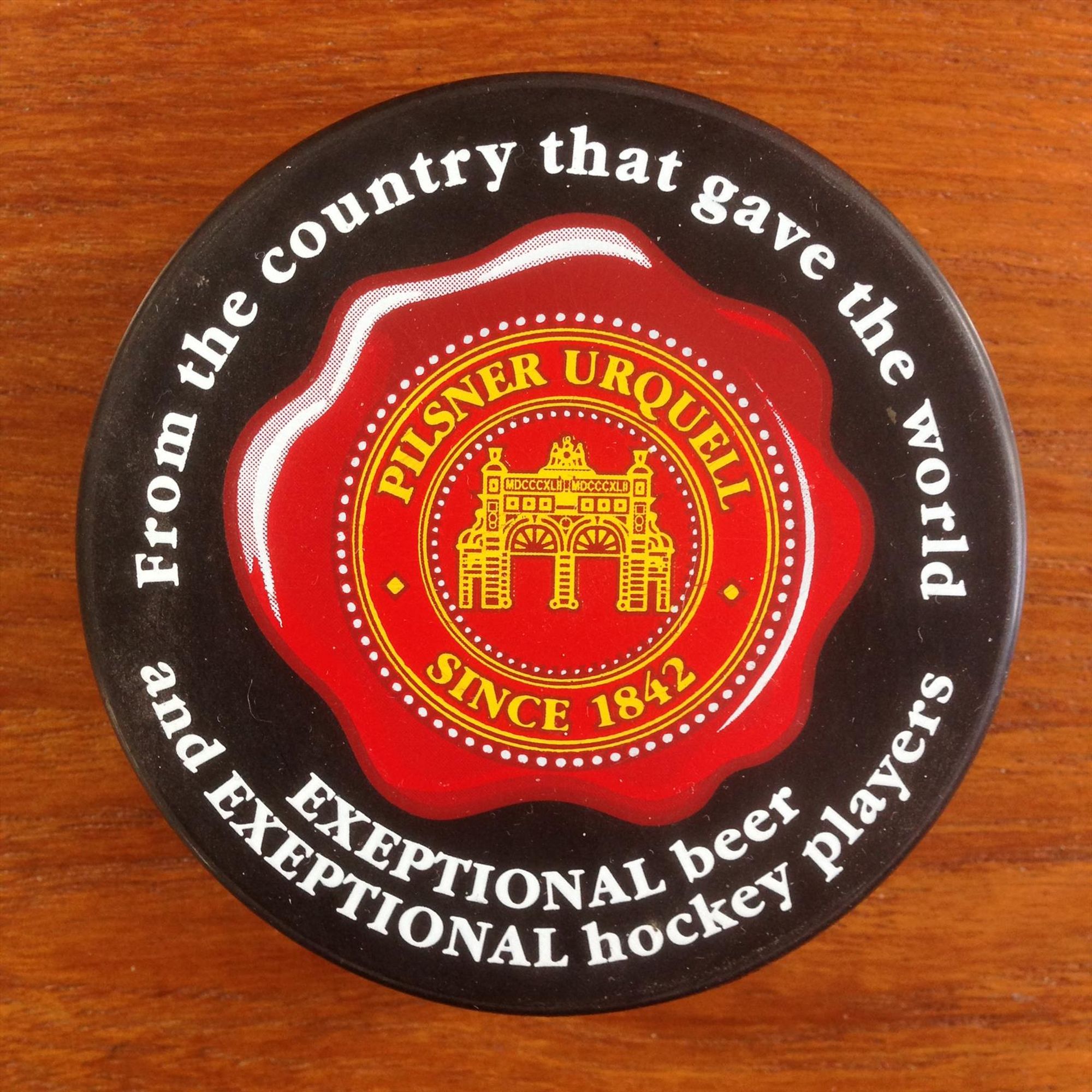 commemorative hockey puck included with cases of Pilsner Urquell in the US

gold-and-red Pilsner Urquell logo in the center

in white around the edge of the black puck:

from the country that gave the world
EXCEPTIONAL beer
and EXCEPTIONAL hockey player