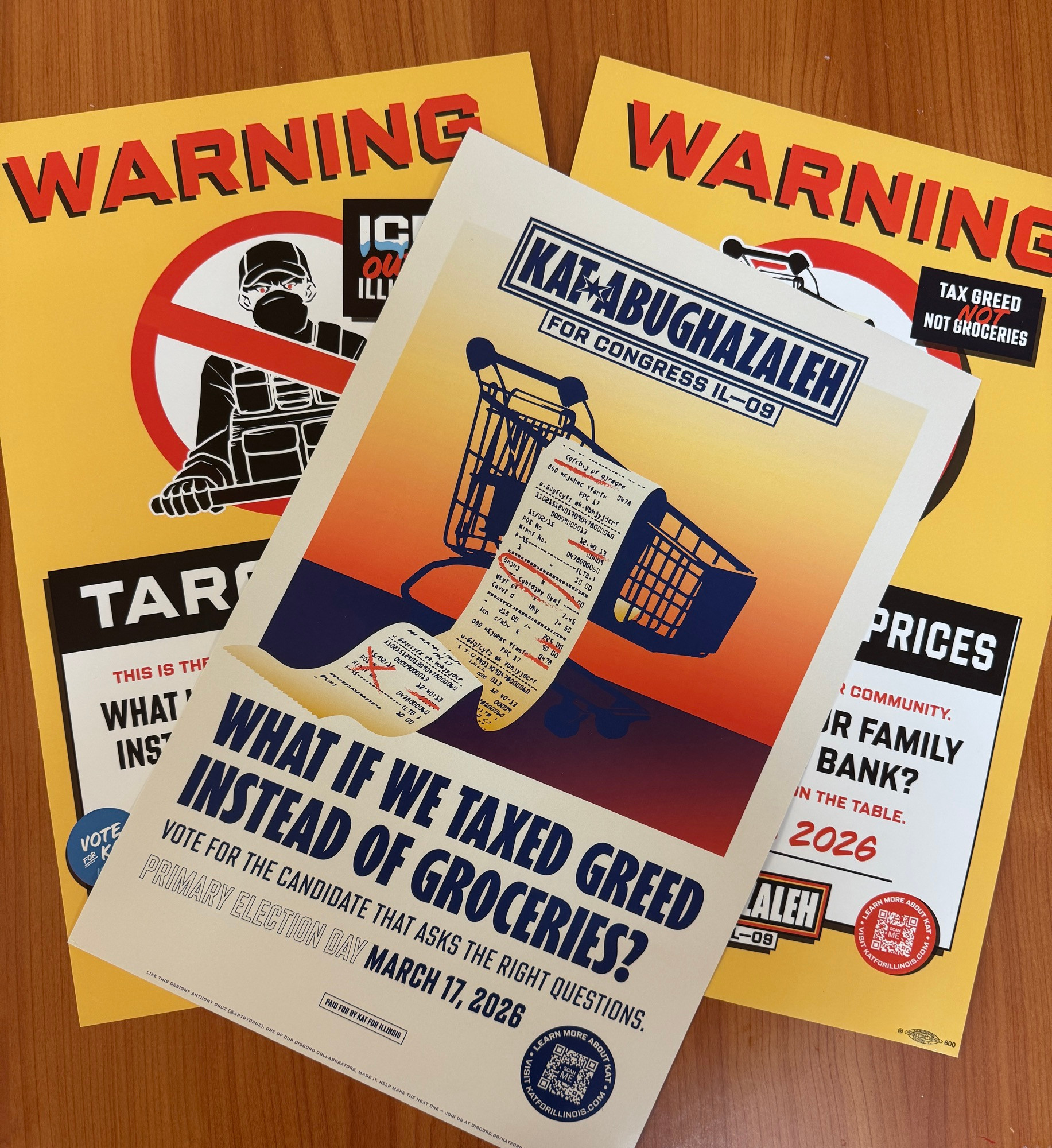 Three posters sit on a brown desk. On top is an ecru poster with an illustration of a grocery cart and an endless receipt marked up with red ink. At the top is the campaign logo and at the bottom in large letters reads: “WHAT IF WE TAXED GREED INSTEAD OF GROCERIES? Vote for the candidate that asks the right questions. PRIMARY ELECTION DAY MARCH 17 2026”

Below are two yellow posters partially obscured. Both say “WARNING” at the top with red circles. The visible circle has a picture of an ICE agent with red eyes and a red line through it.