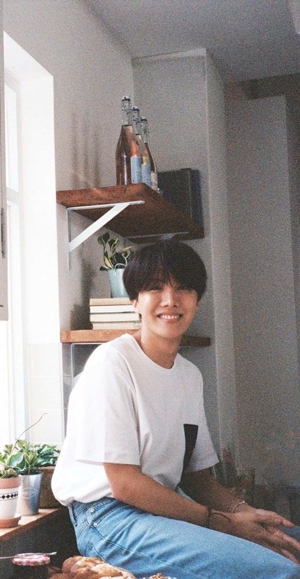 This is a picture of J-hope in white t-shirt and blue jeans sitting by a wooden shelf with plants and bottles.