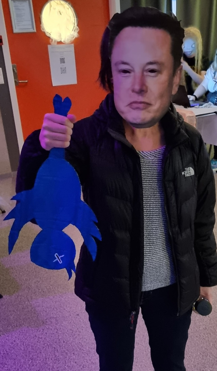 A person wearing a mask which is simply Elon Musk's face printed on paper, and holding a dead, blue bird cut out from cardboard by its feet. The bird's dead eyes have crosses over them resembling the X logo for Twitter.