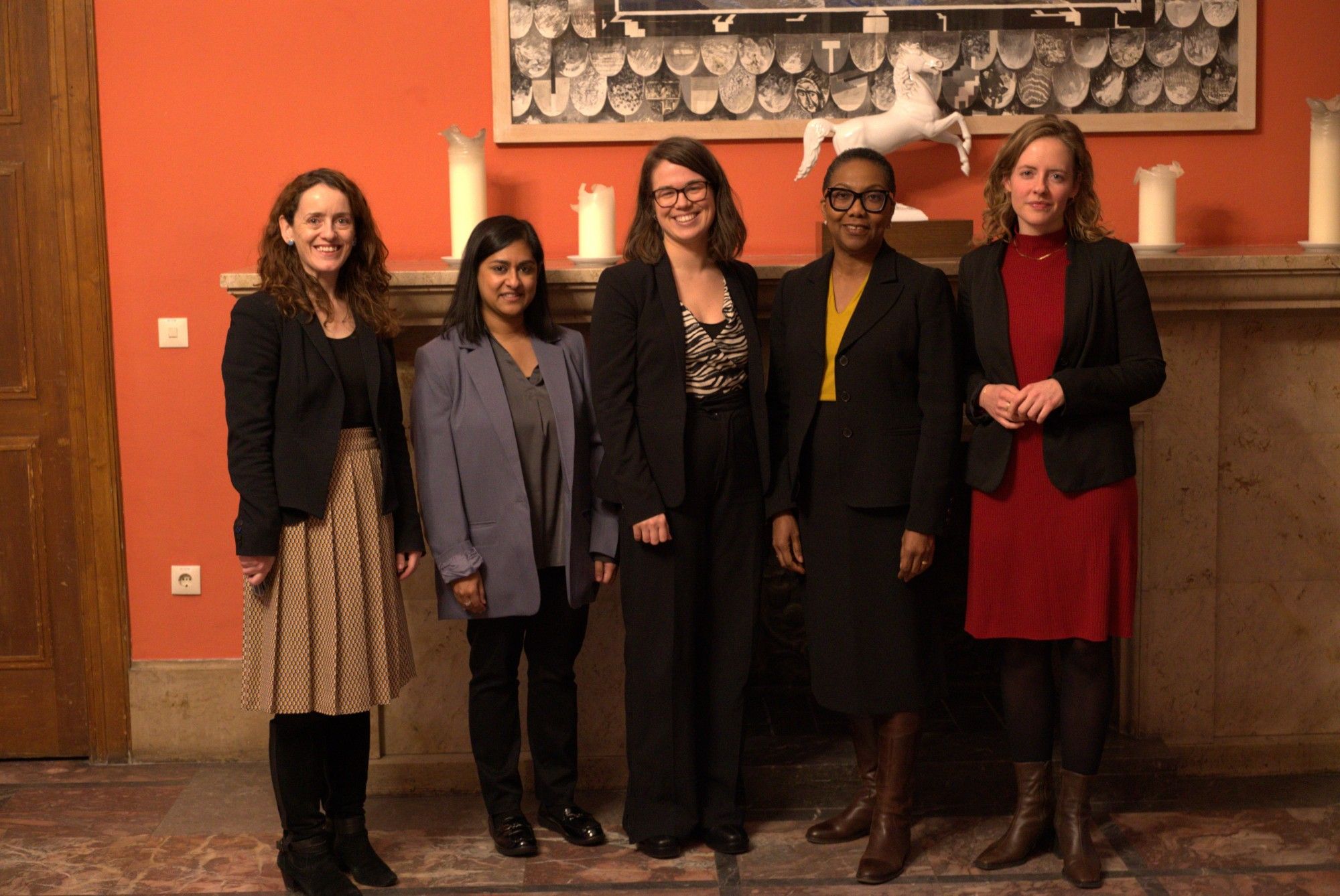 A group of 5 women, amongst them our guest Alicia Herbert, Research Fellow Leonie Stamm & Research Assistant Anna Hauschild, is standing in front of an orange wall and a chimney.