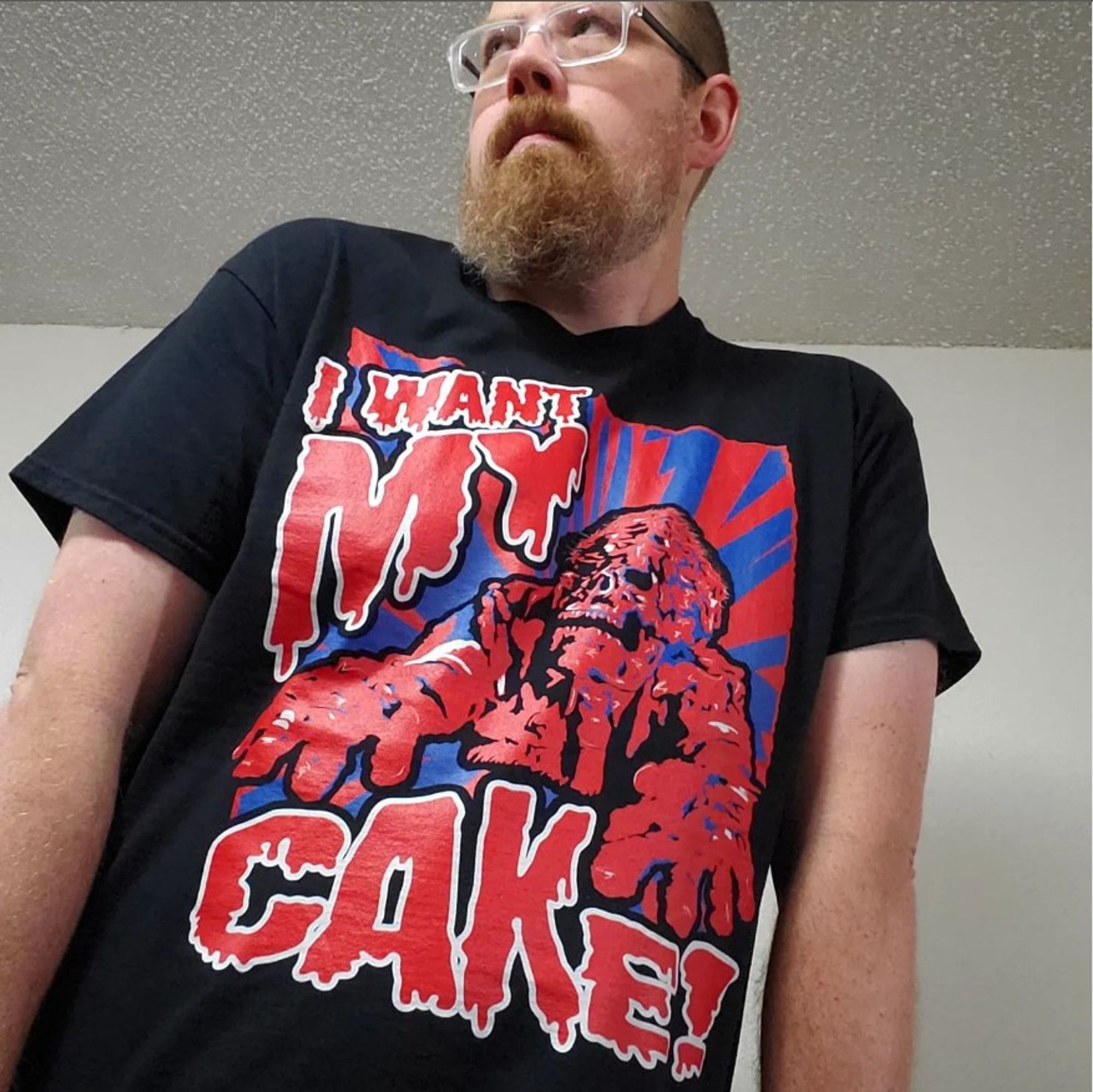 A man in glasses wearing a black t-shirt with a red and blue zombie illustration on it that reads "I want my cake!"