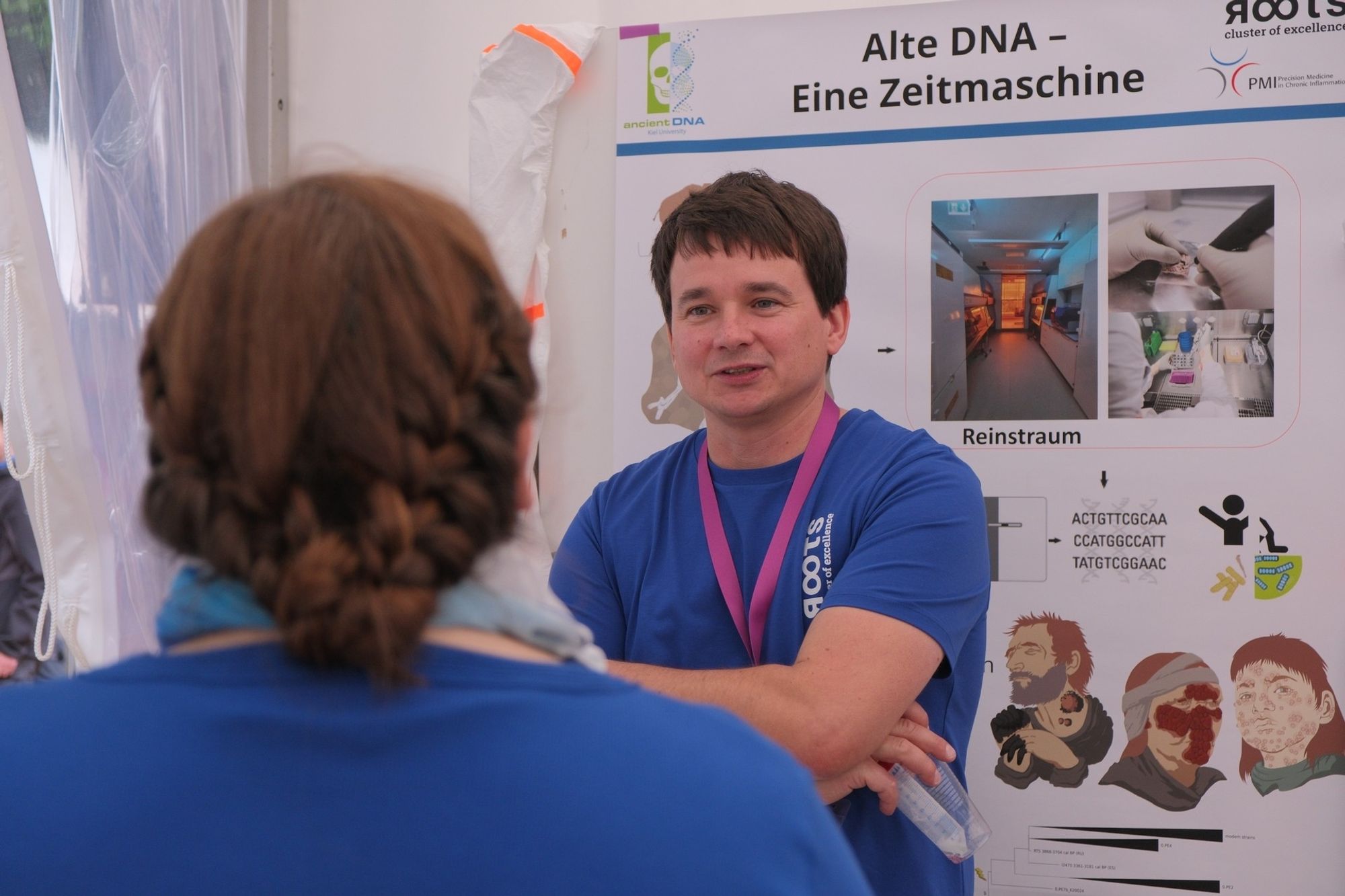 Eine Person steht vor einem Poster, auf dem "Alte DNA - Eine Zeitmaschine" steht und unterhält sich mit einer zweiten Person, die von hinten zu sehen ist.