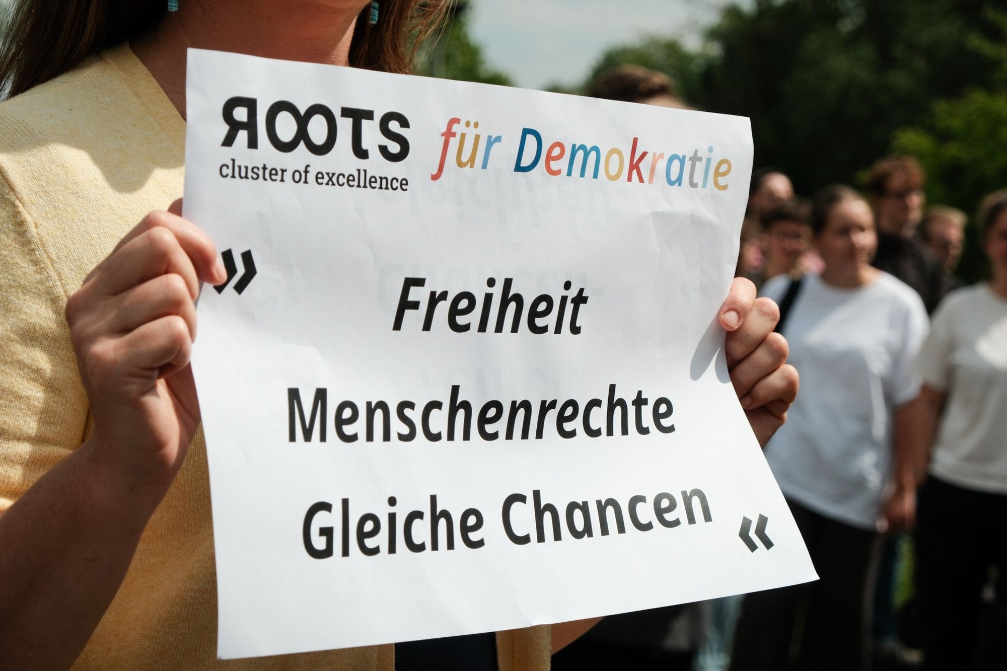 In einer Menschenmenge hält eine Person ein Blatt mit der Aufschrift "ROOTS für Demokratie - Freiheit, Menschenrechte, Gleiche Chancen" hoch.