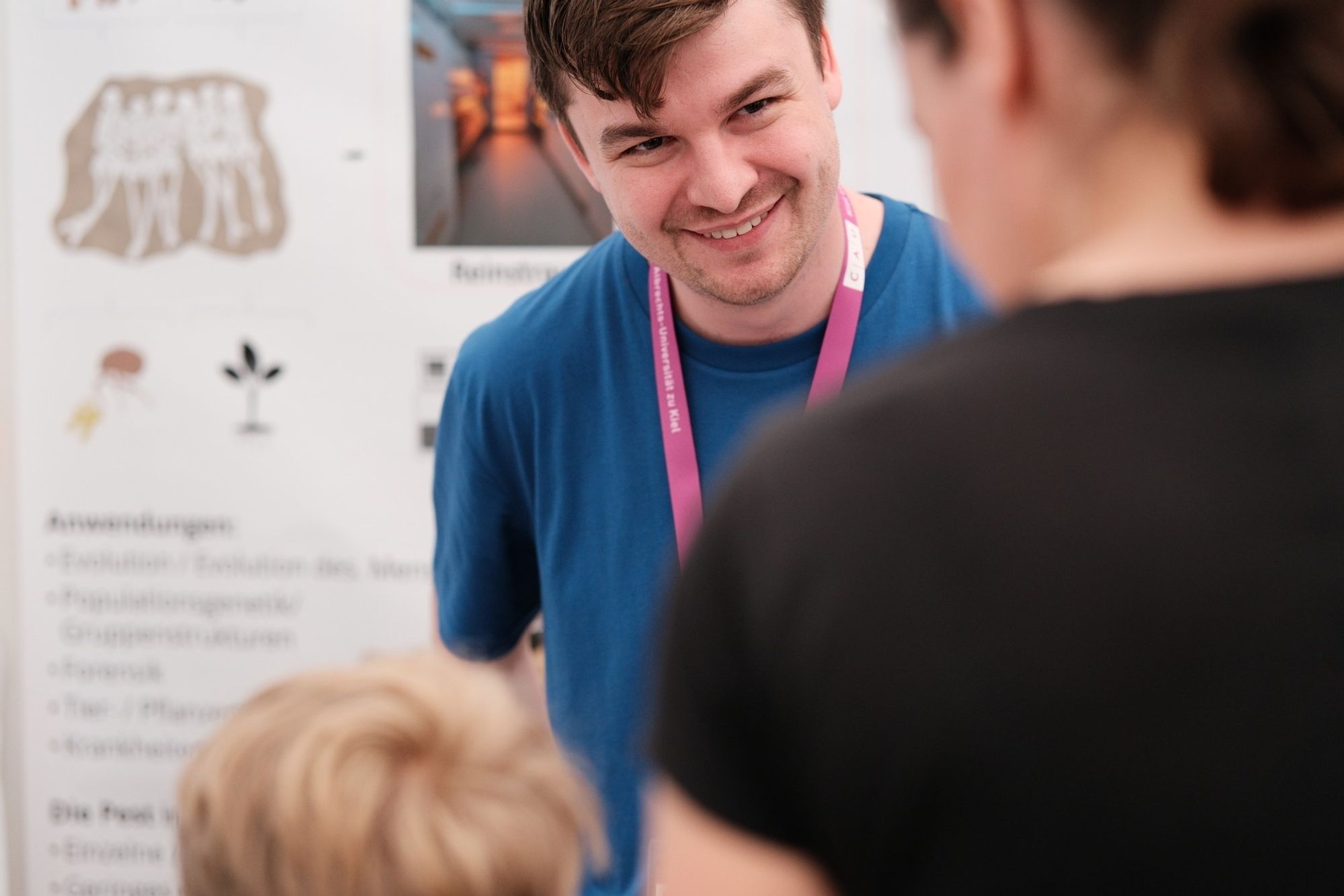 Ein Person vor einem Poster lächelt eine erwachsene Person und ein Kind an, die mit dem Rücken zur Kamera stehen