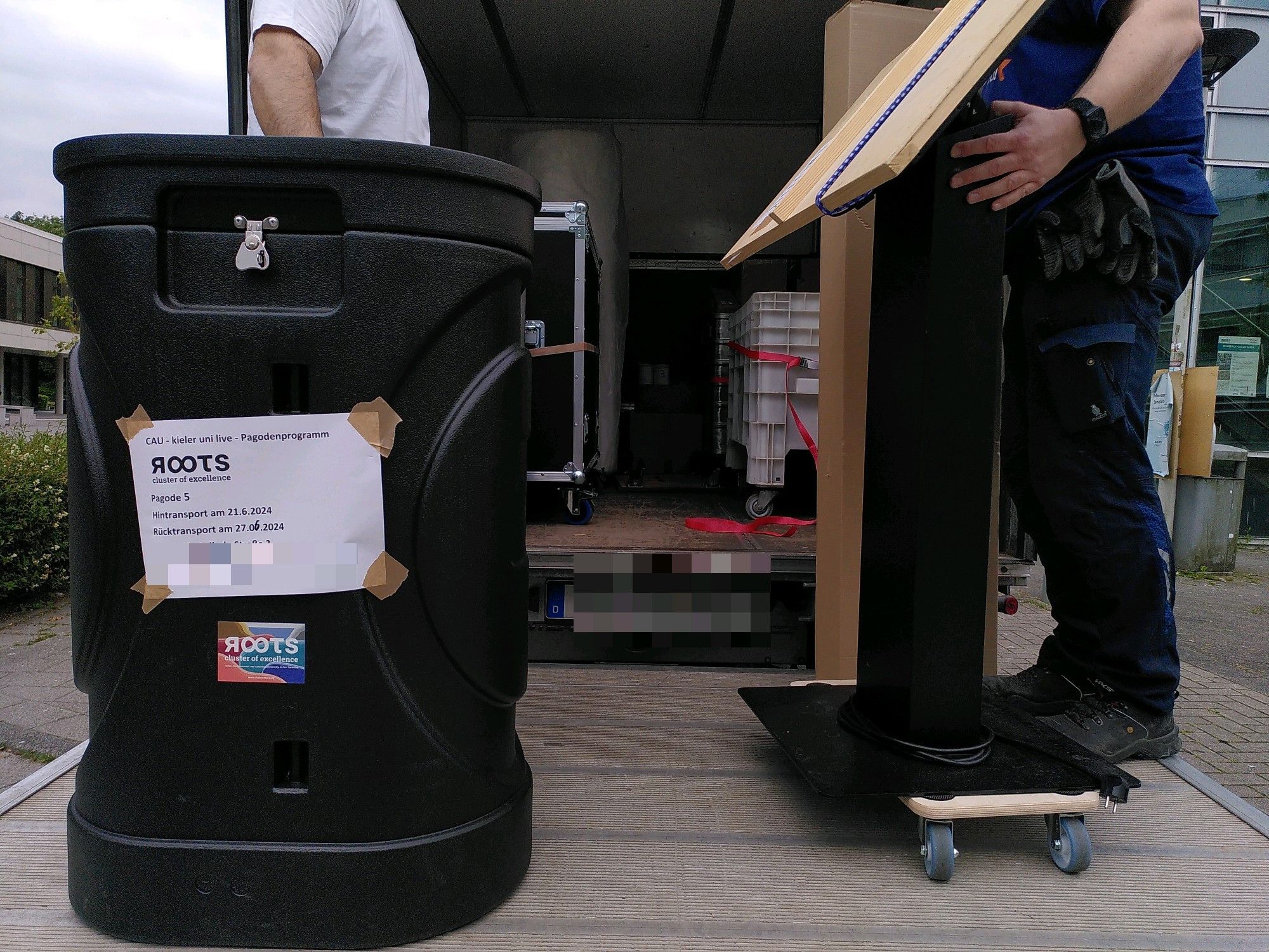 Eine rollbare Tonne und ein verpackter Bildschirm auf der Hebebühne eines Transporter. Auf der Ladefläche sind bereits mehrere Kisten und Boxen.