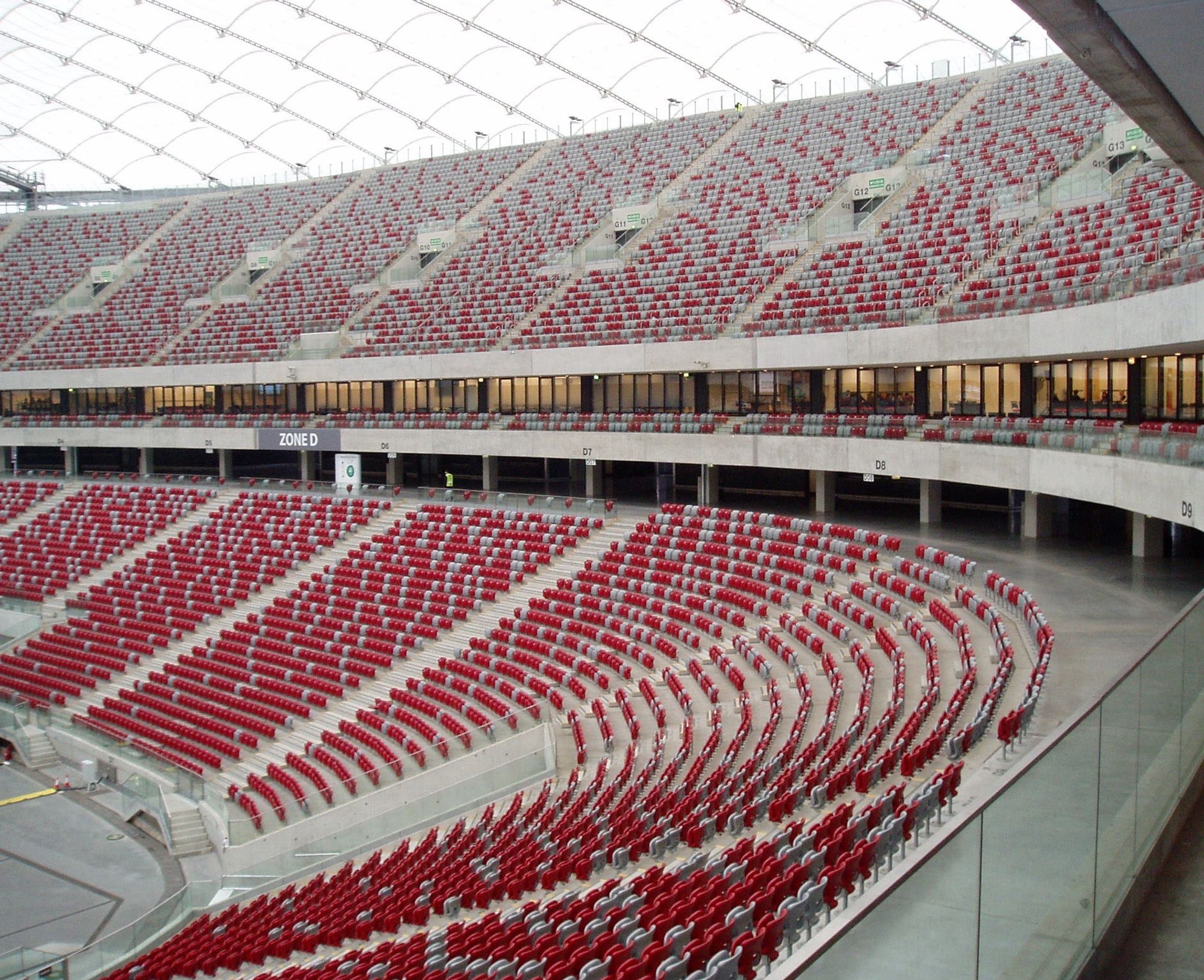 Rot-weiße Sitzschalen in einem Fußballstadion, verteilt auf zwei Ränge, halbrund angeordnet und unterbrochen durch eine Ebene mit Logen. Man sieht viel grauen Beton.