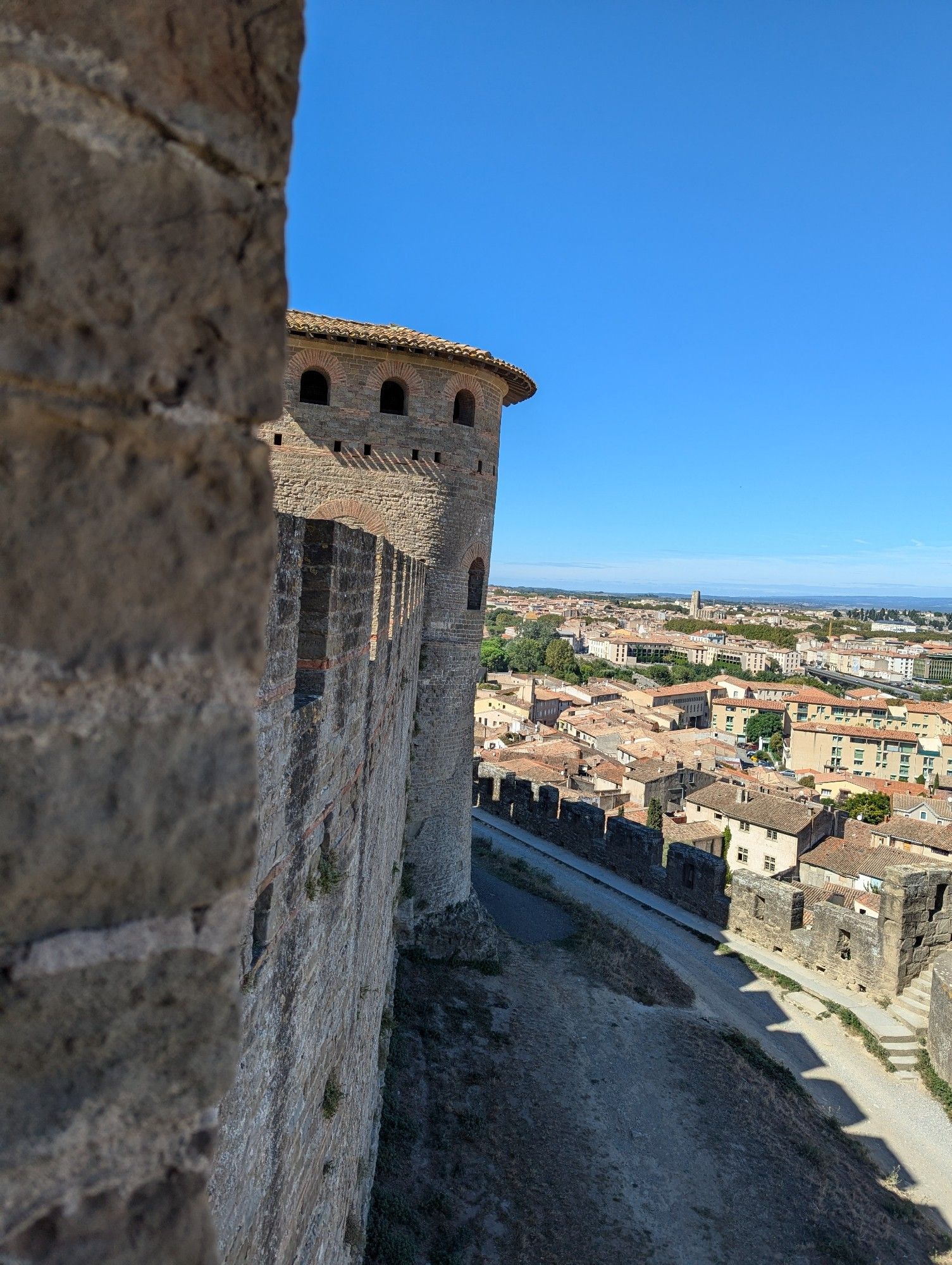 Looking along a stone wall towards a tower, with a lower rampart and town below. The sky is blue