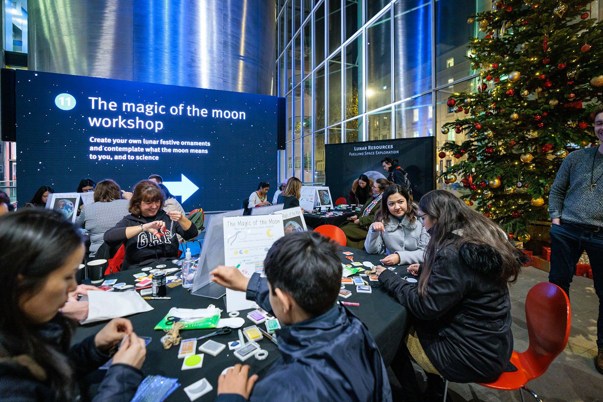 People sit around tables with lots of art materials, behind them is a sign saying 'The magic of the moon workshop' and a Christmas tree