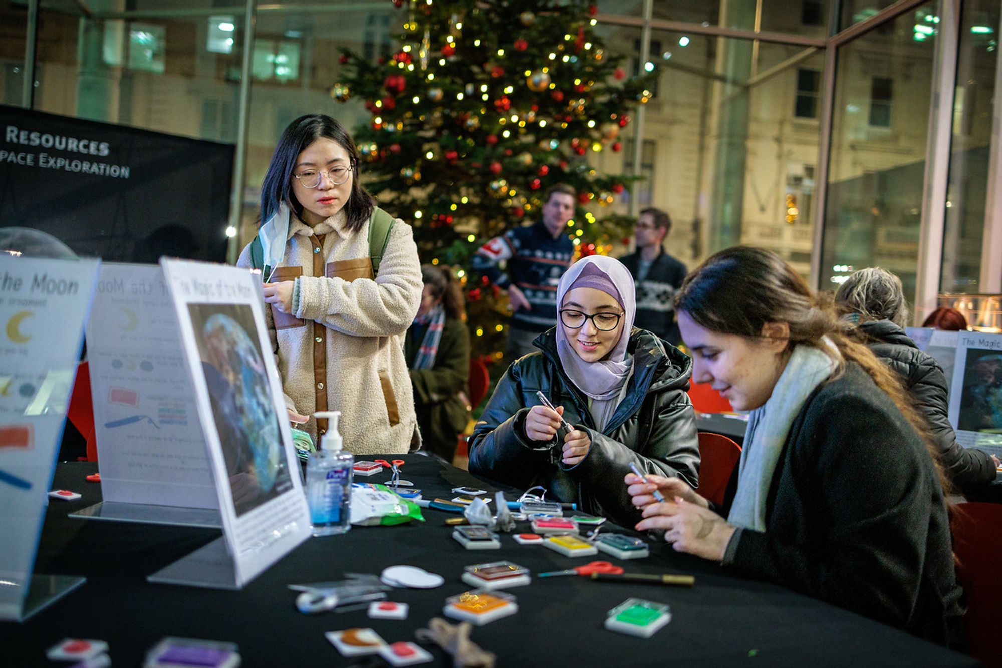 2 people sit at a table crafting while another looks over them. In the background is a Christmas tree