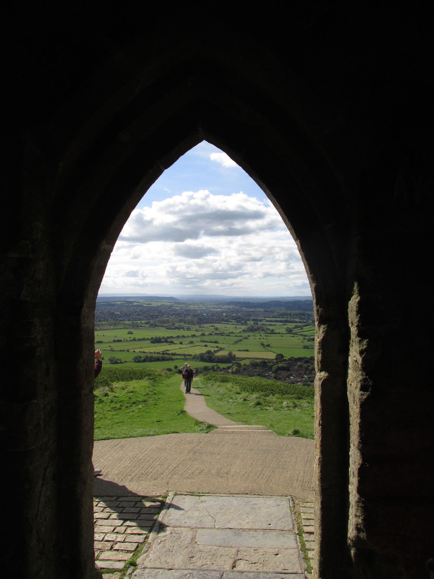 A view through an archway of St Michael's Tower, Glastonbury Tor, looking South-West. Green fields of the Somerset flats along with residential areas are visible under mostly cloudy skies.