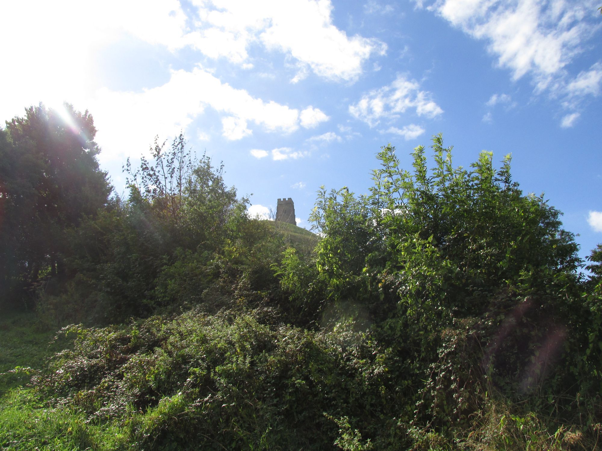 Looking up at St Michael's Tower from part way up Glastonbury Tor, with partly cloudy blue skies, sunlight in the top right and green foliage in the bottom of the frame.