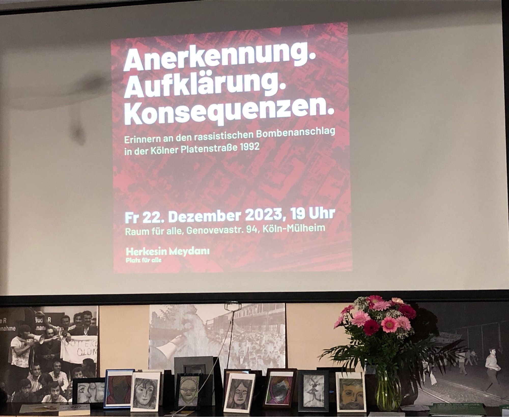 Hintergrund des Veranstaltungsraumes „Raum für Alle“ in Köln Mülheim. Ein Bild auf der Leinwand mit Schrift „Anerkennung. Aufklärung. Konsequenzen. Erinnern an den rassistischen Bombenanschlag in der Kölner Platenstraße 1992“. Davor Gemälde und Zeichnungen des Künstlers Ali Rıza Ceylan, Blumen.