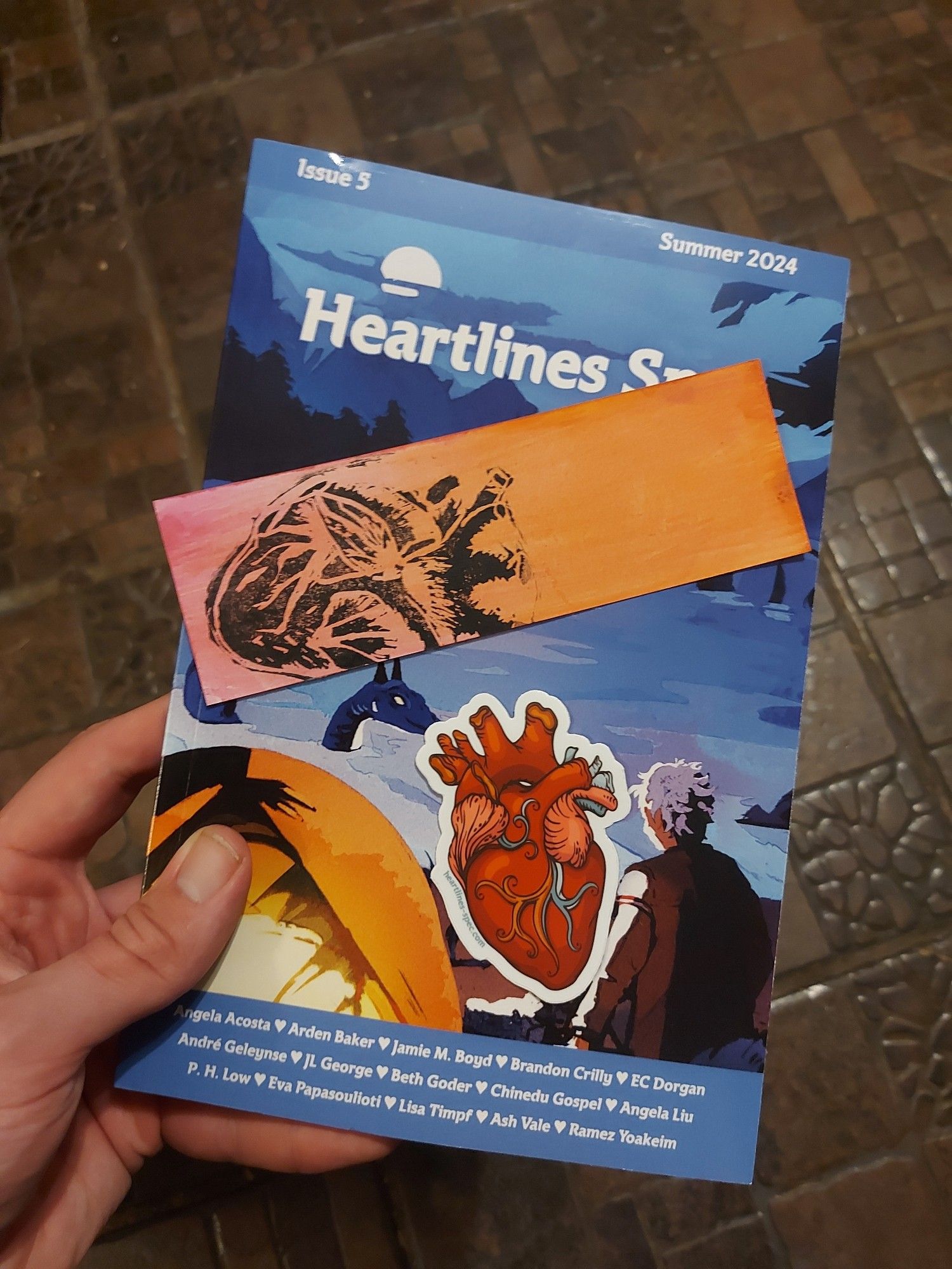 My hand holding the paperback of Heartlines Spec Issue 5. Balanced on it are a bookmark with an anatomically accurate heart stamp (the Heartlines logo) and a Heartlines logo sticker.