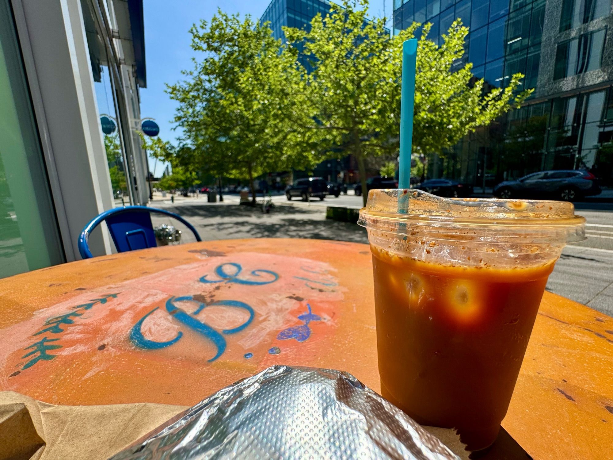 Outside on the sunny sidewalk of a city street, a bright-orange table with the letters “BC dc” stenciled on it in blue. Atop the table are an iced coffee and a foil-wrapped sandwich. In the distance beyond, a bright green tree.