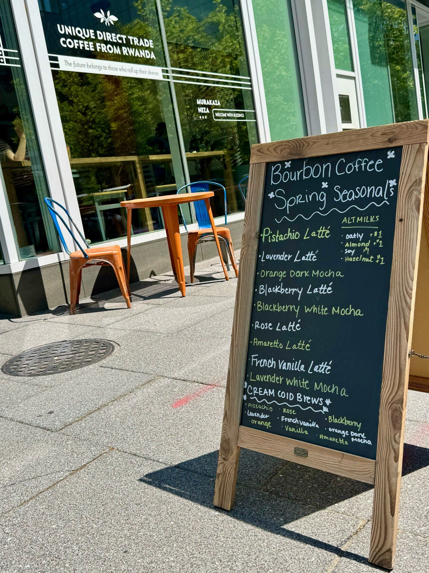 Sandwich board on a sunny sidewalk, a black chalkboard with a list of fancy coffee drinks under the heading “Bourbon Coffee Spring Seasonals.” Beyond, a set of two chairs and a table, all painted in gaudy orange with blue accents. Beyond that, the front of Bourbon Coffee, a shop advertising “unique direct trade coffee from Rwanda.”