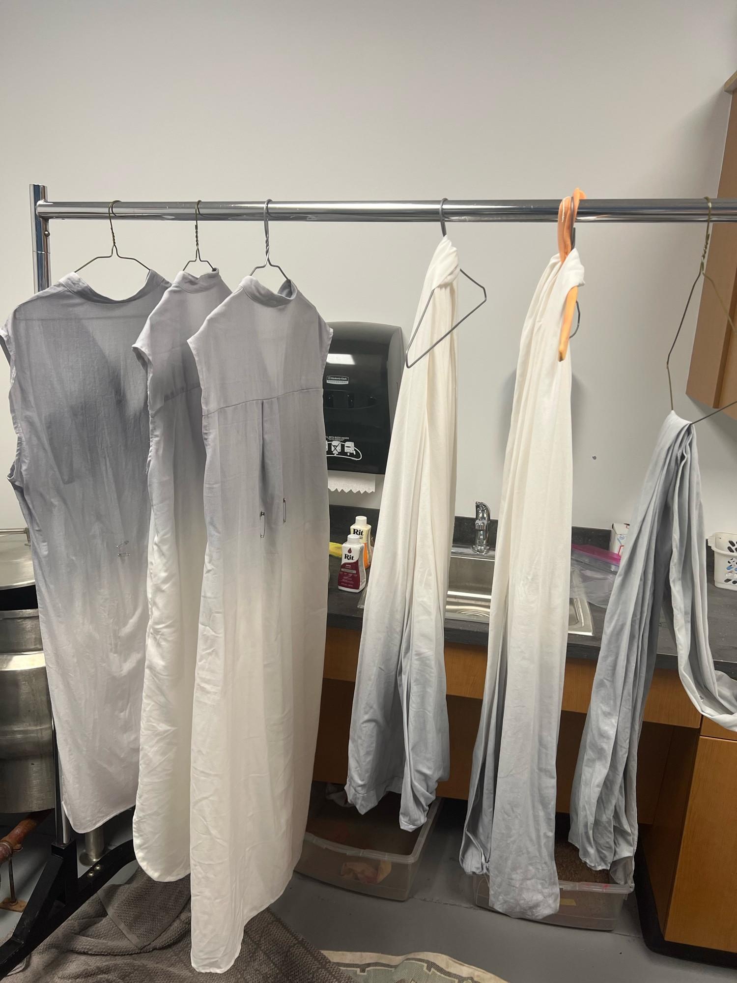 Three tunic dressses and three drapes of linen fabric hanging on a z rack infront of a sink and dye vat. The dresses and fabrics have a medium grey tone that transitions into white