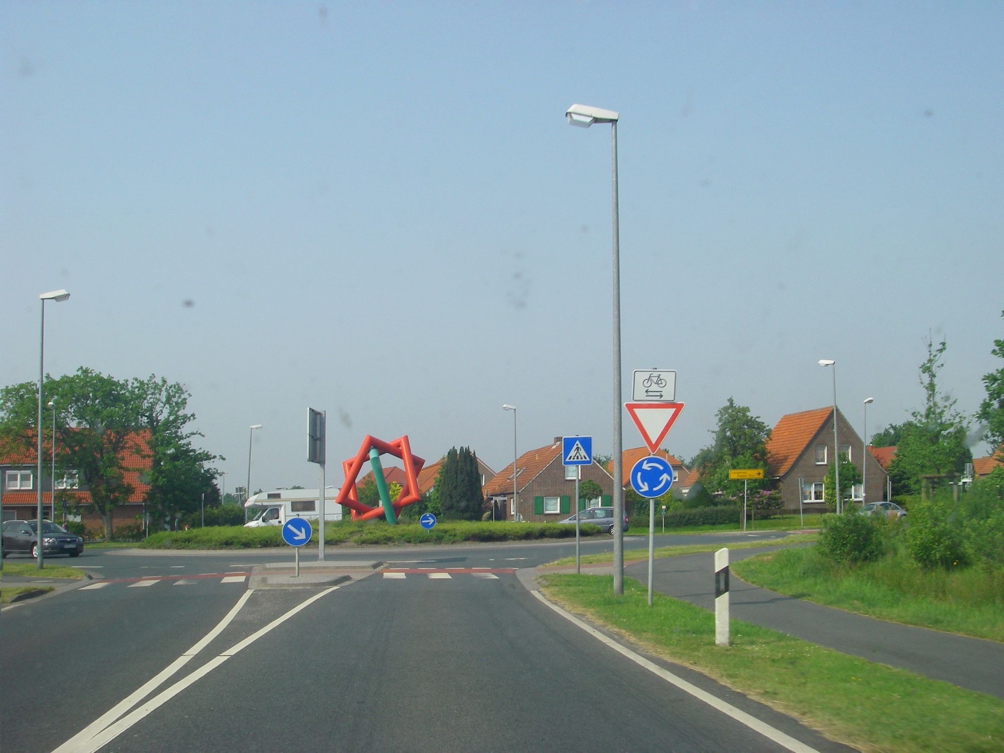 Straße, die auf einen Kreisverkehr zu führt. In der Mitte des Kreisverkehrs eine Skulptur aus drei in einander verschachtelten Quadraten die aus Rohren geformt sind.