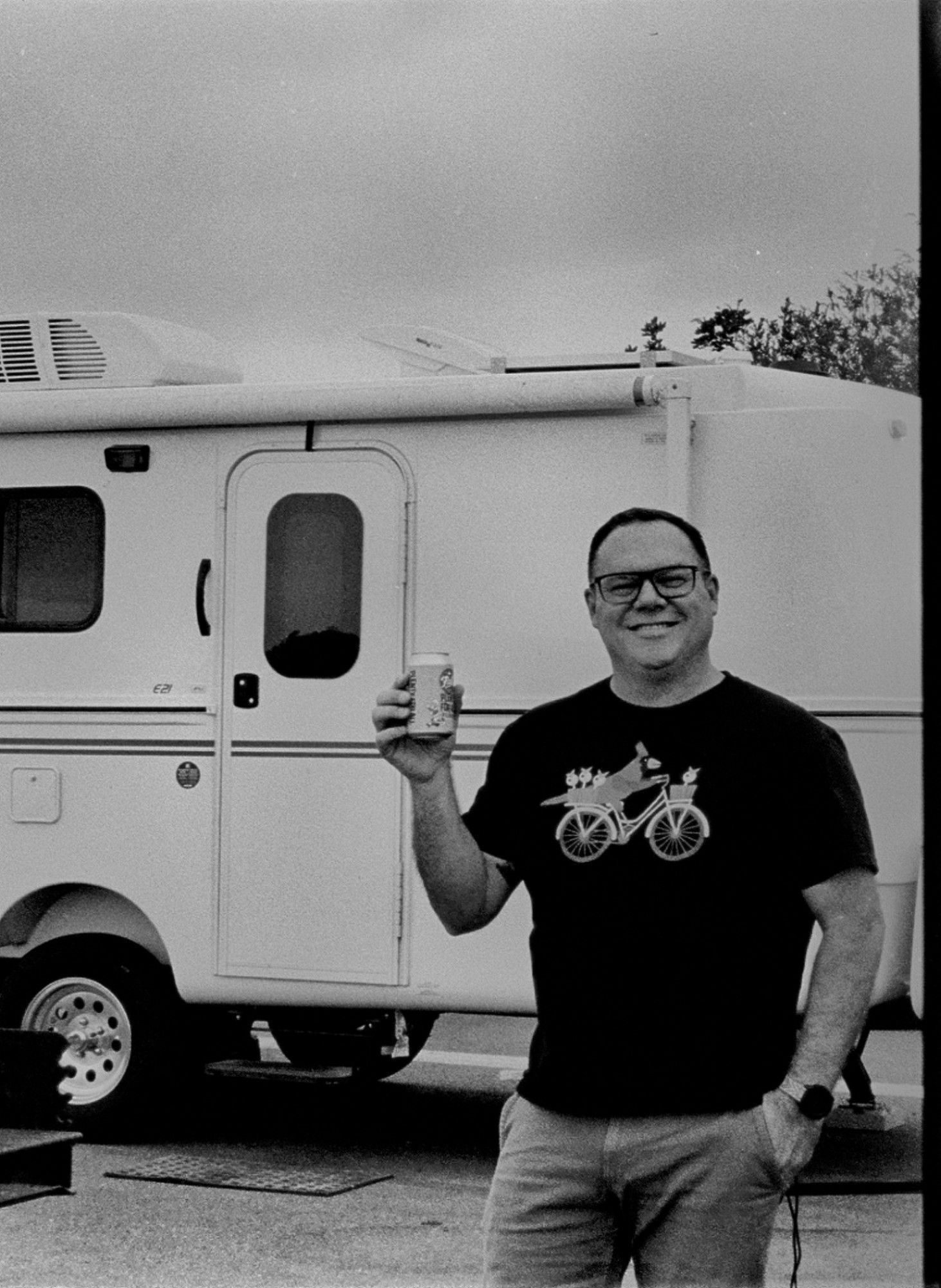 A black and white picture of some dude (me) in a black shirt with a bird riding a bicycle in front of a travel trailer. He has a beer in one hand, the other hand is in his pocket