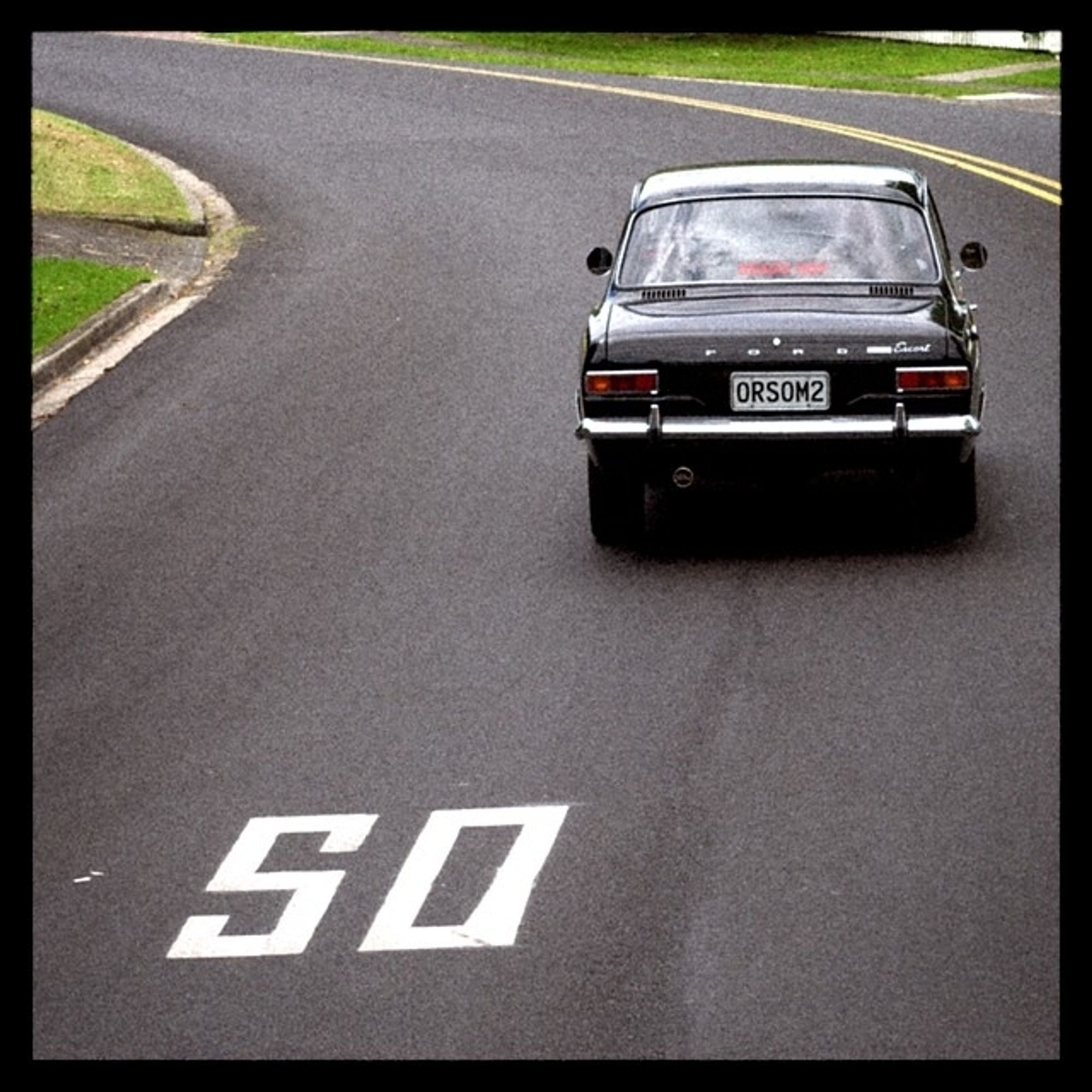 An old black ford escort drives away up a hill. The number 50 is painted in large figures on the road. The 5 looks like an S. The licence plate of the car reads "ORSOM".
Tauranga, New Zealand.