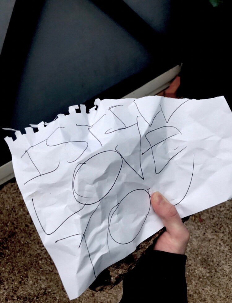 A ripped piece of paper that says “I STILL LOVE YOU” in messy handwriting, wrapped around a rock. Picture taken in ominous location.