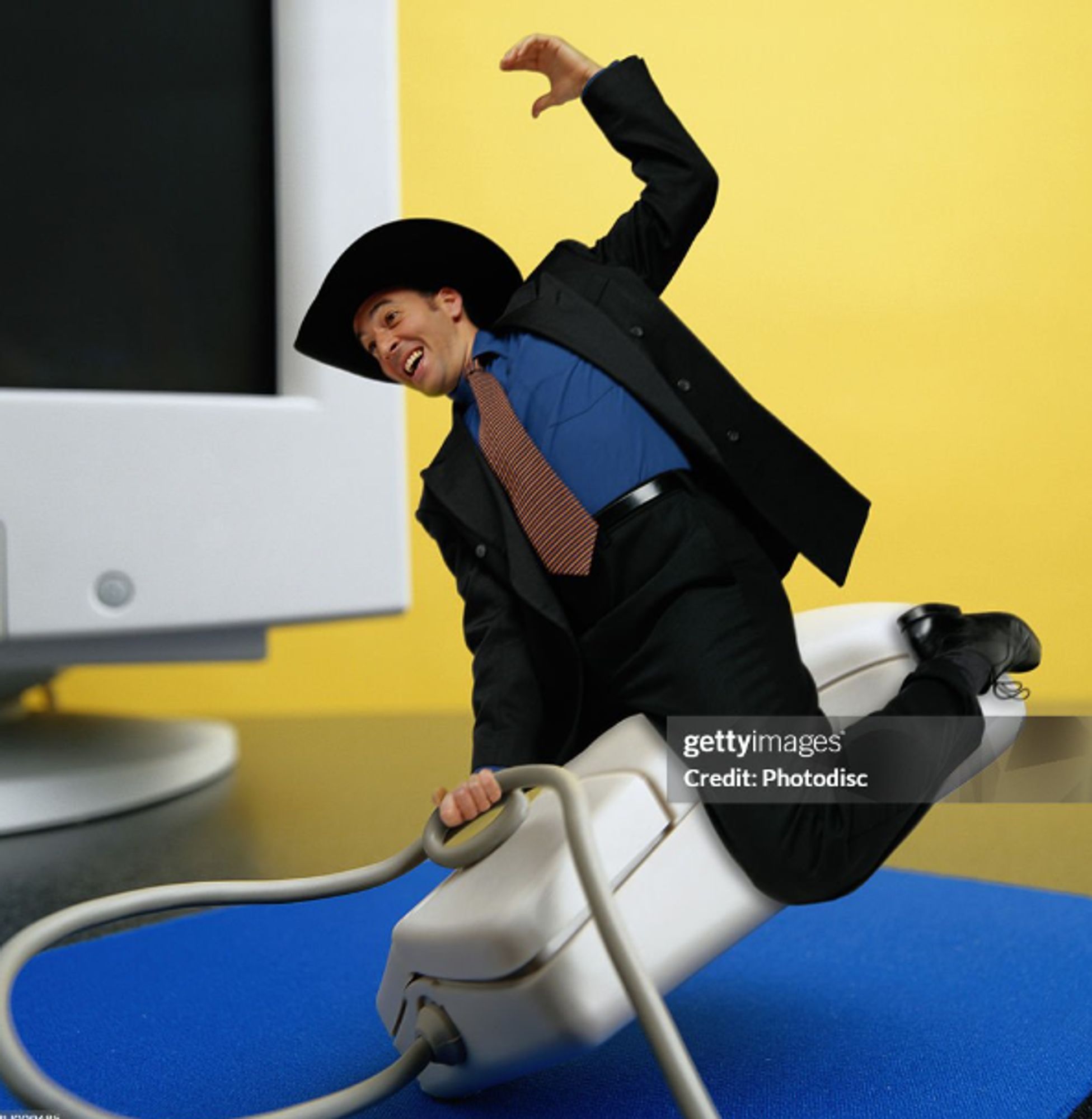 a screenshot of a getty stock photo of a white man in a black suit and tie wearing a black cowboy hat while riding astride a giant computer mouse like it's a bucking bronco