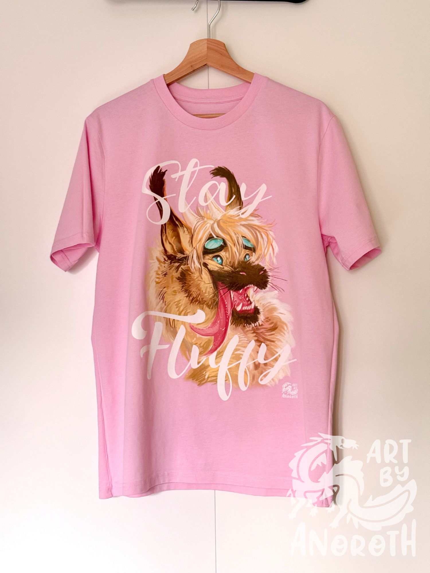 a pastel pink tshirt with a cute fluffy furry beige dragon smiling , around the dragon portrait wraps around text saying: "Stay fluffy"