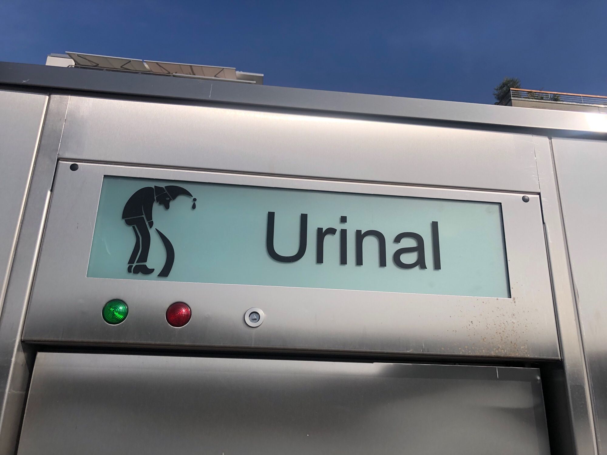 Schild über einer öffentlichen Toilette, Aufschrift „Urinal“, Piktogramm zeigt Mann mit Zipfelmütze in nach vorne gebeugter Haltung, er schaut hinab auf seinen dicken Urinstrahl