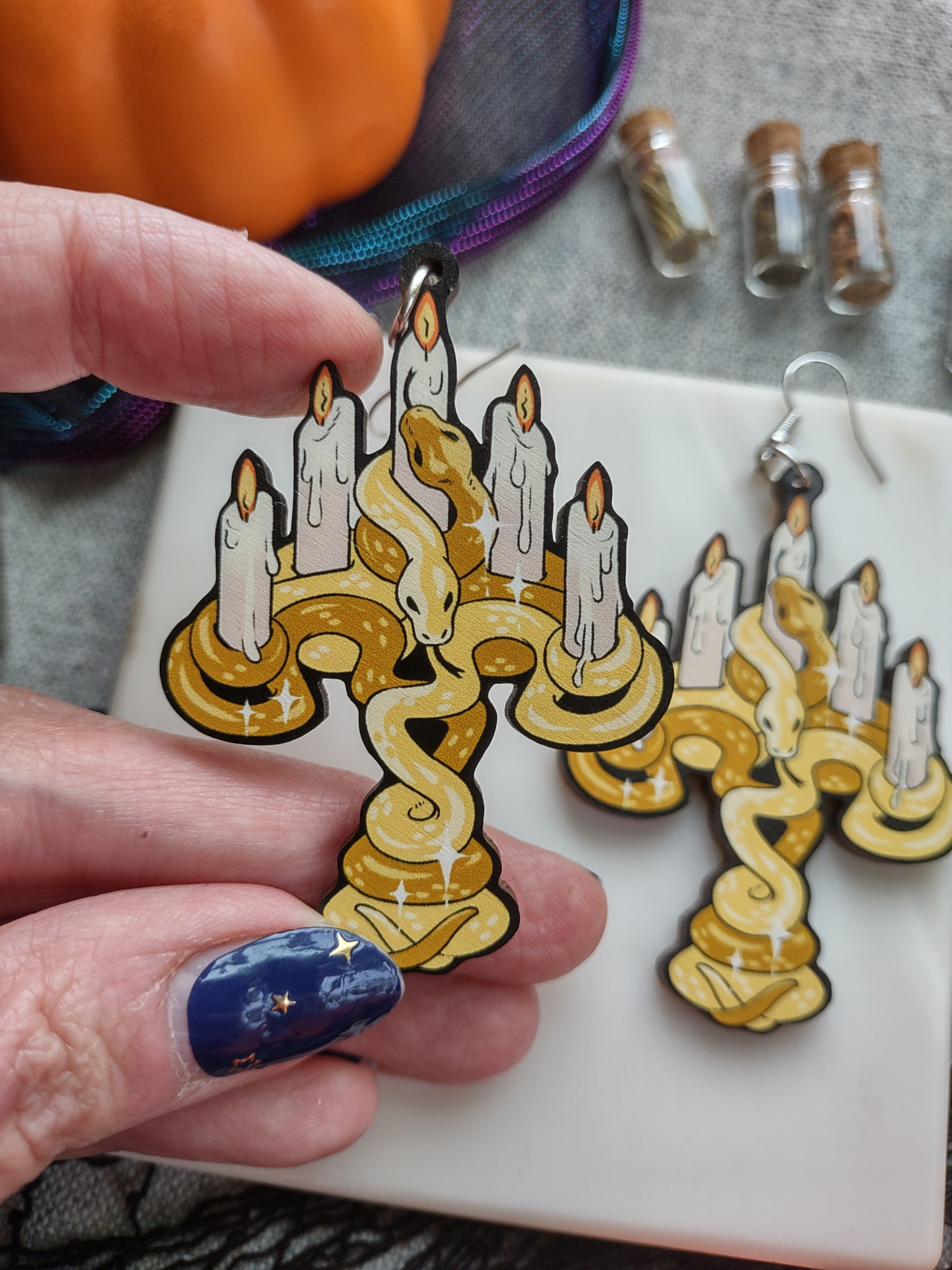 A pair of wooden earrings featuring golden colored snakes wrapped around each other in the shape of a candelabra. The candelabra holds 5 candles and the two snakes faces rest at the base of the 5th candle in the center.