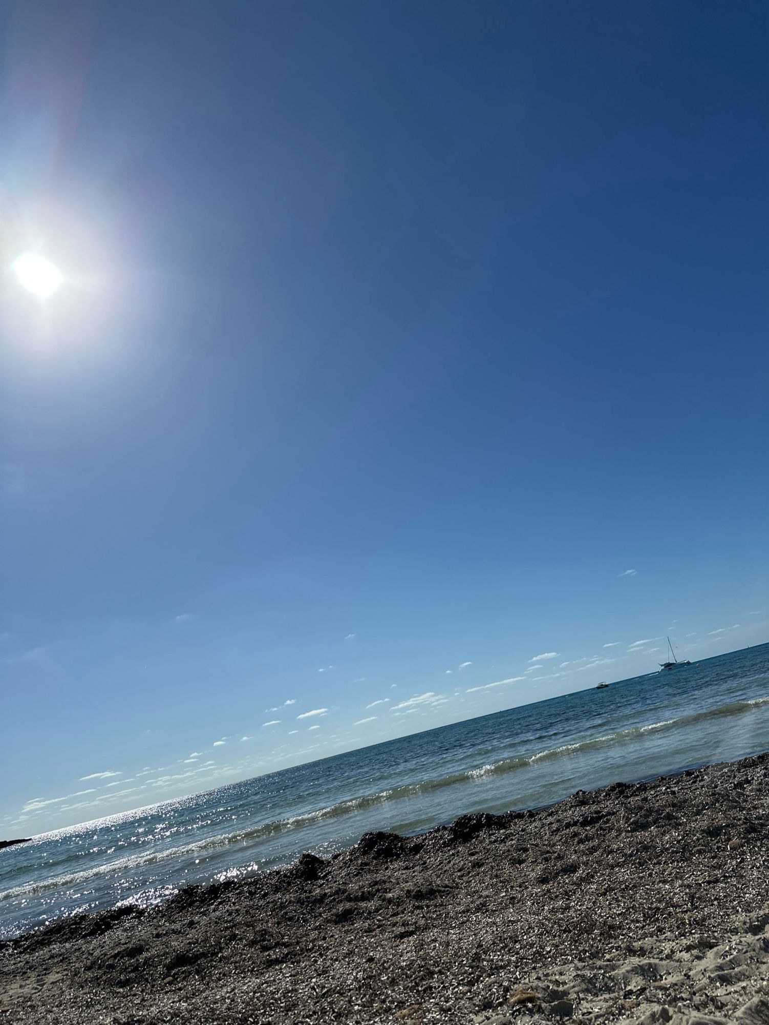 POV am Strand liegend, Sand mit Seegras und leichte Wellen. Himmel blau mit wenigen Wolken.