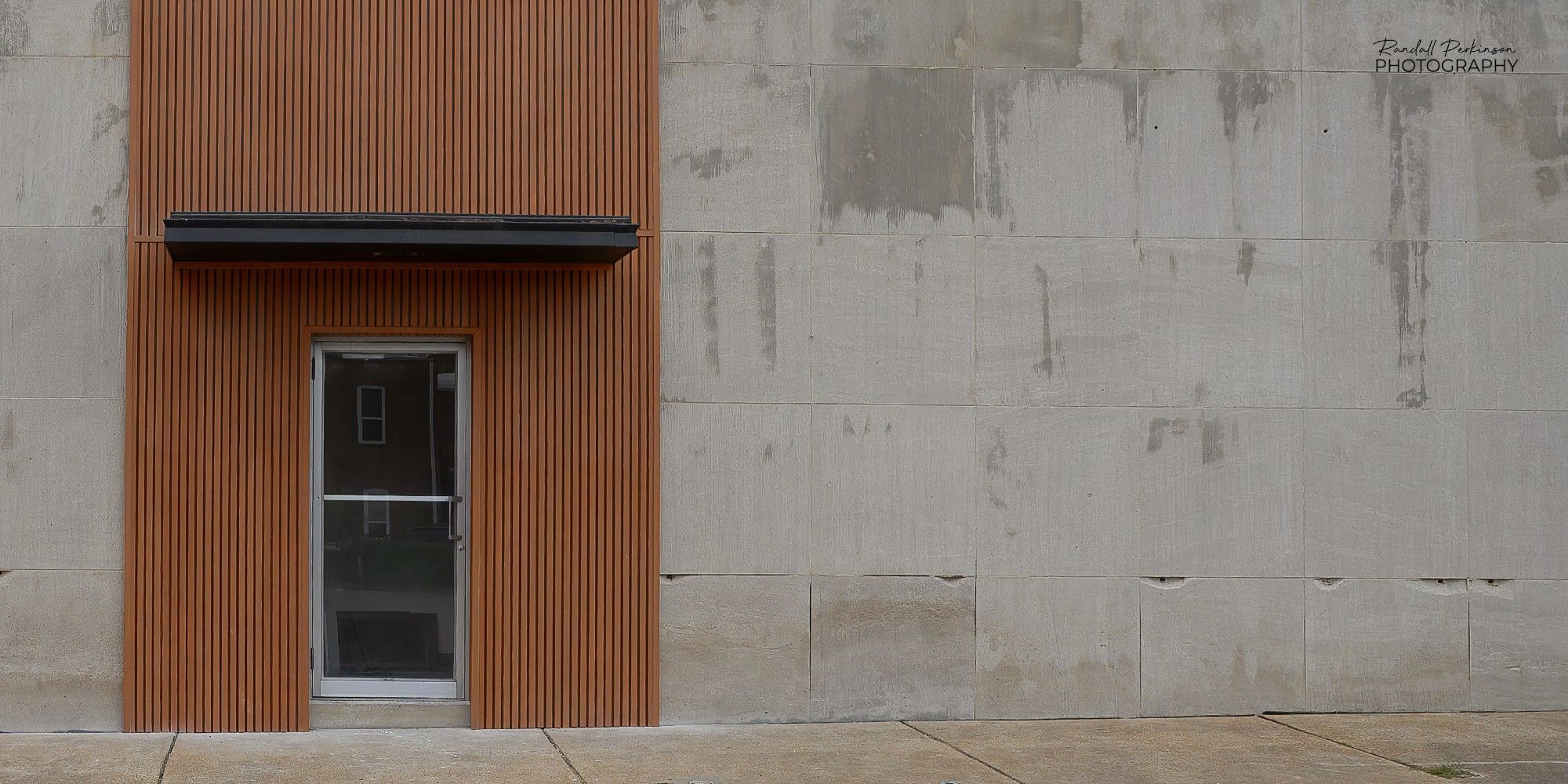 A single metal and glass door entrance in a concrete building is embellished with vertical brown wood slats for a few feet either side of the door and a small black awning over the door.