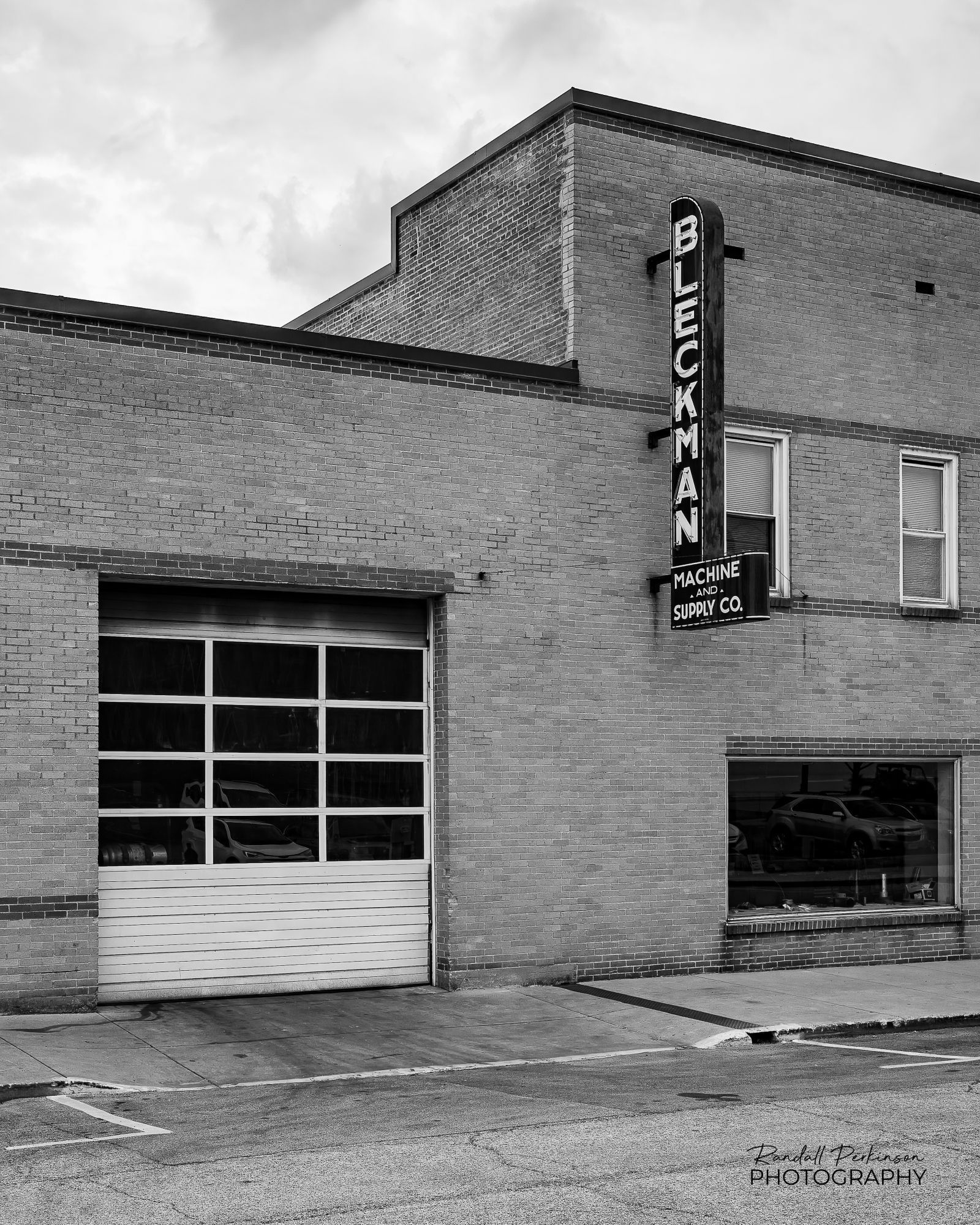 A vintage sign with "Bleckman" appearing vertically and "Machine and Supply Co" appearing in smaller letters horizontally hangs on the side of a two story brick building with a large garage door.