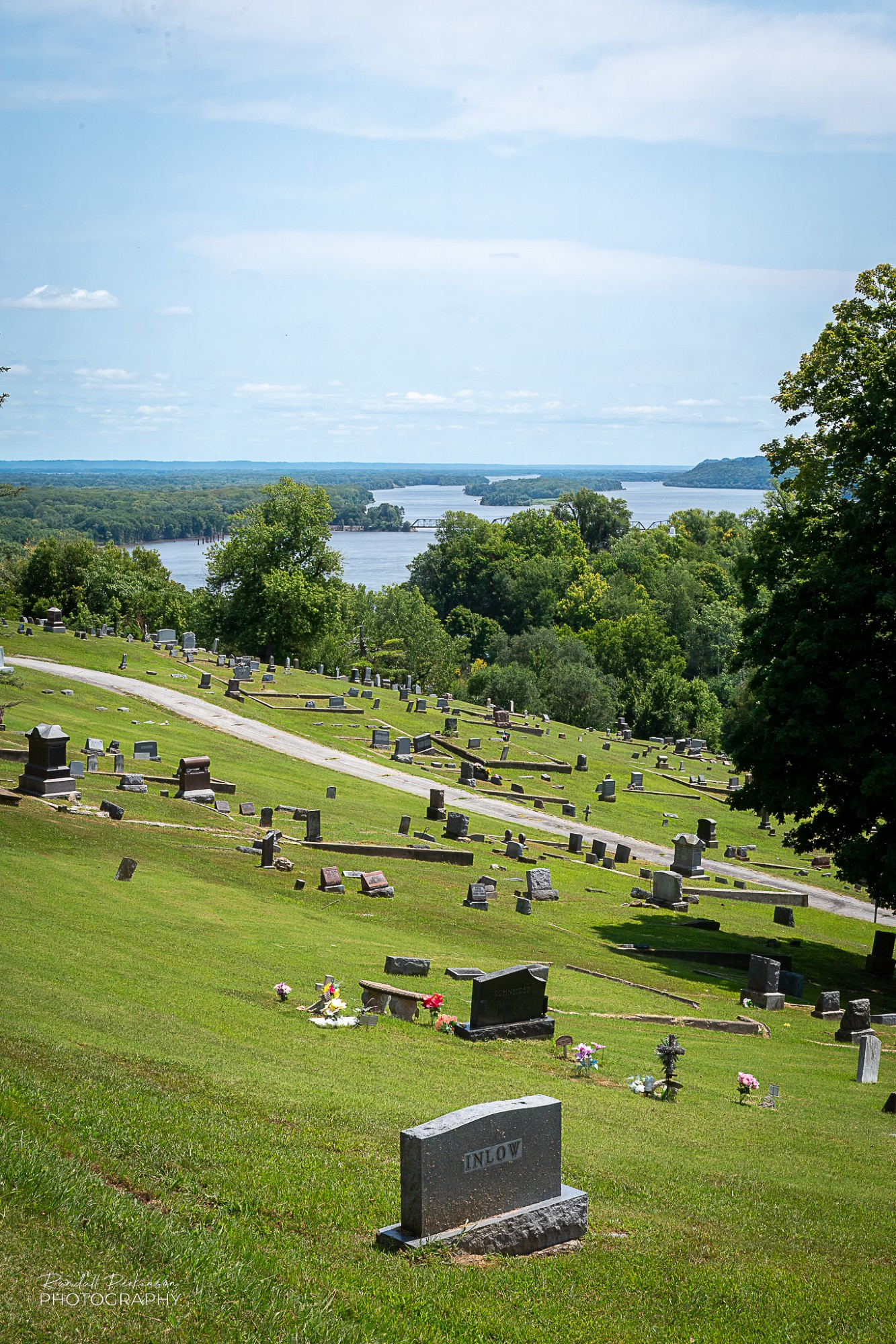 Gravestones are spread across a hillside cemetery with views of the Mississippi River in the background.