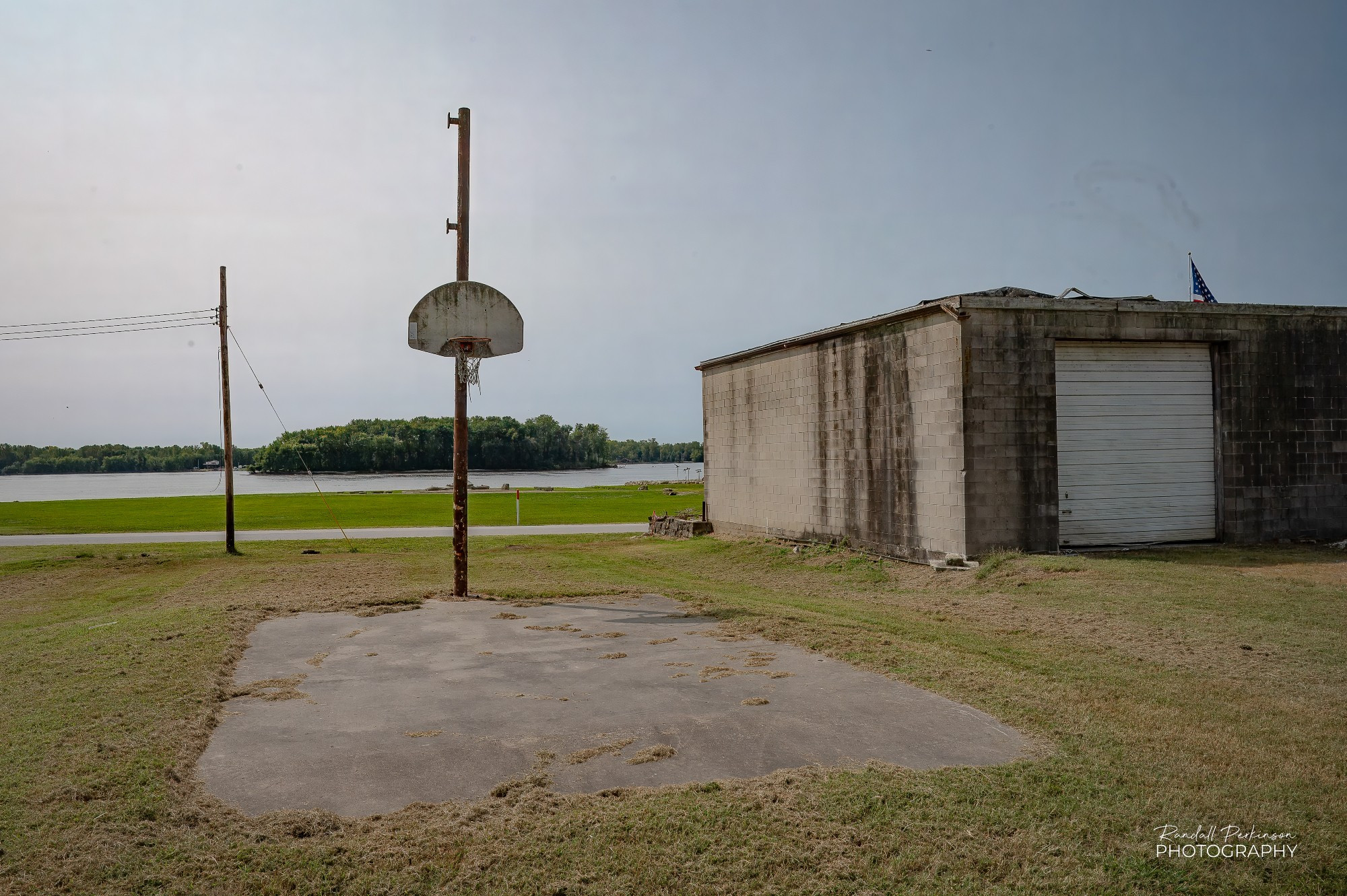 A basketball goal on a wood power pole sits at the end of a small paved area near an old single story concrete block building.  The Mississippi River runs in the background with forested banks on the opposite side.