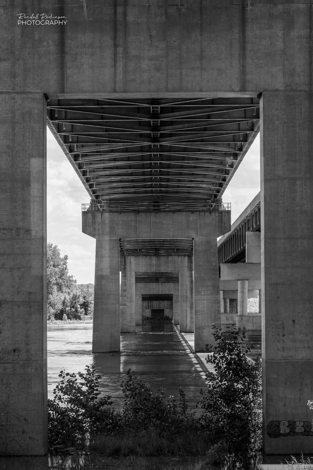 Looking along the length of a large steel truss river bridge from below the bridge and between the legs of the concrete piers.