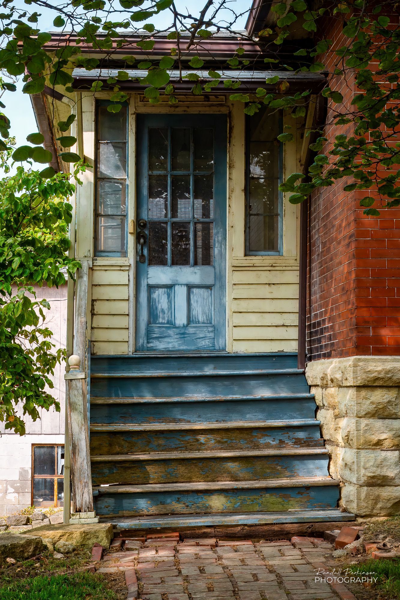 A faded blue door and six steps down to a brick sidewalk.  The door is on a framed addition at the back of a brick building with a stone foundation.